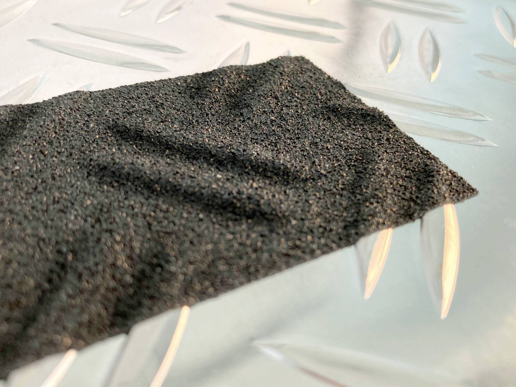 Schwarzes Sandpapierstück liegt auf einer silbernen Metallplatte mit Rautenmuster.