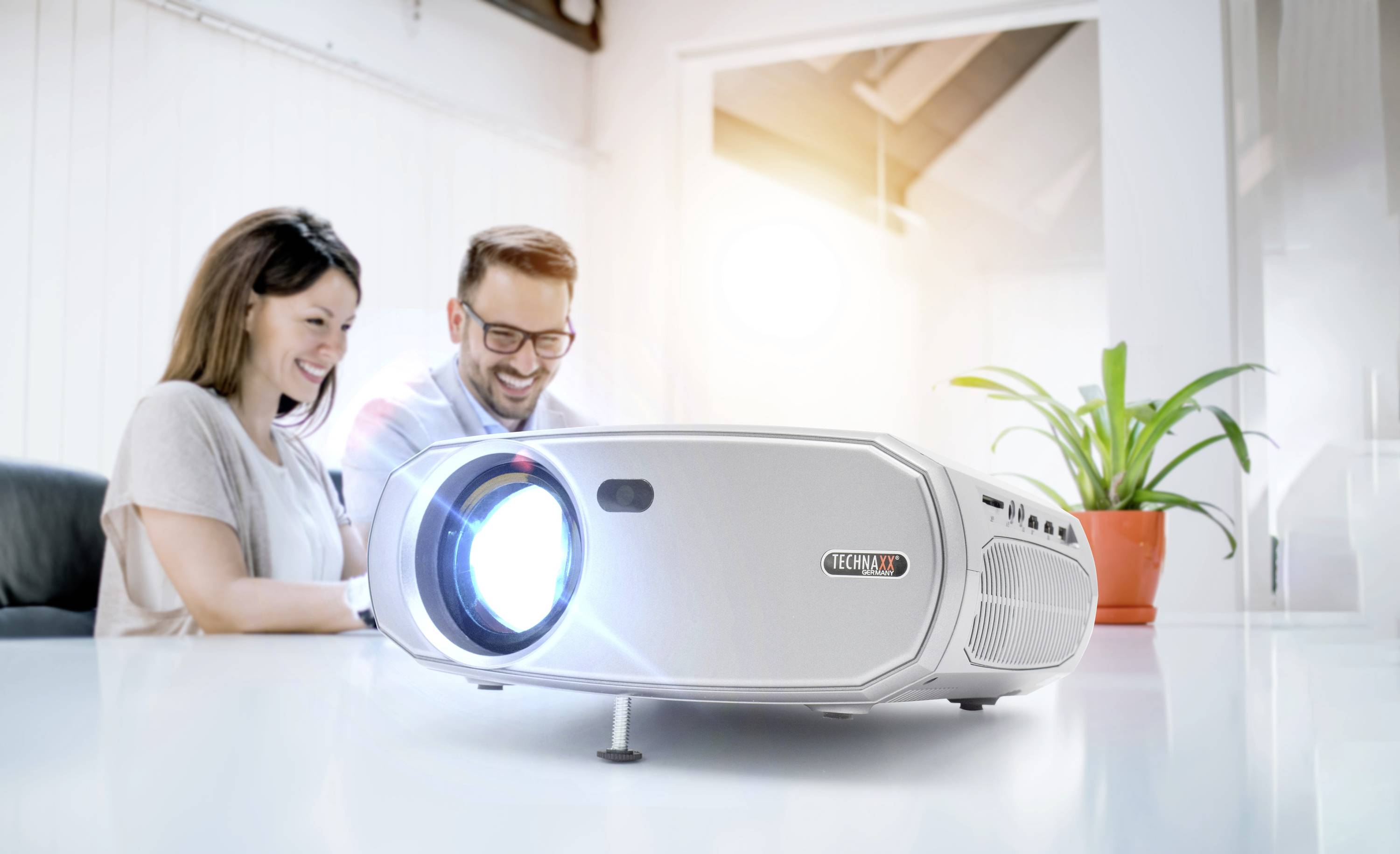 Ein Projektor strahlt ein helles Licht auf einem Tisch. Im Hintergrund sitzen zwei Personen lächelnd nebeneinander bei einem Meeting.