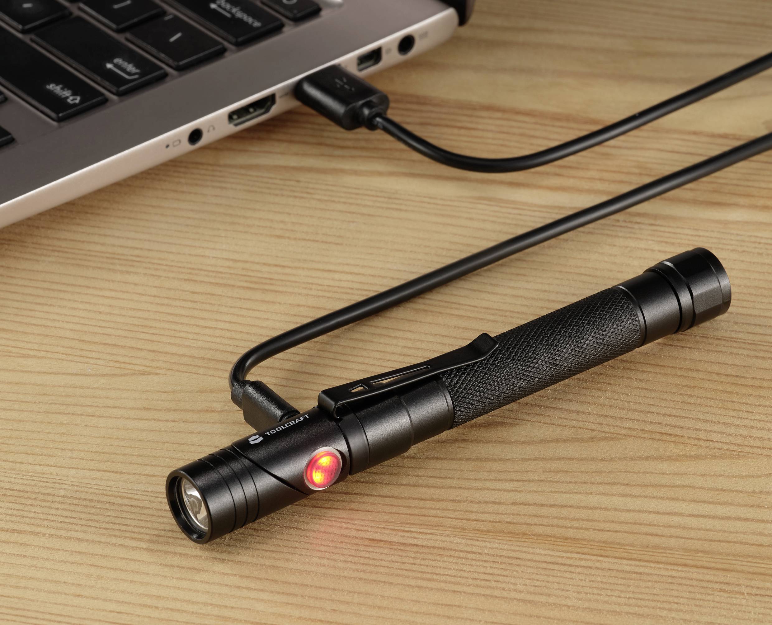 Schwarze Taschenlampe liegt auf holzfarbenem Tisch, angeschlossen an Laptop über USB-Kabel, rotes Licht an der Taschenlampe leuchtet.