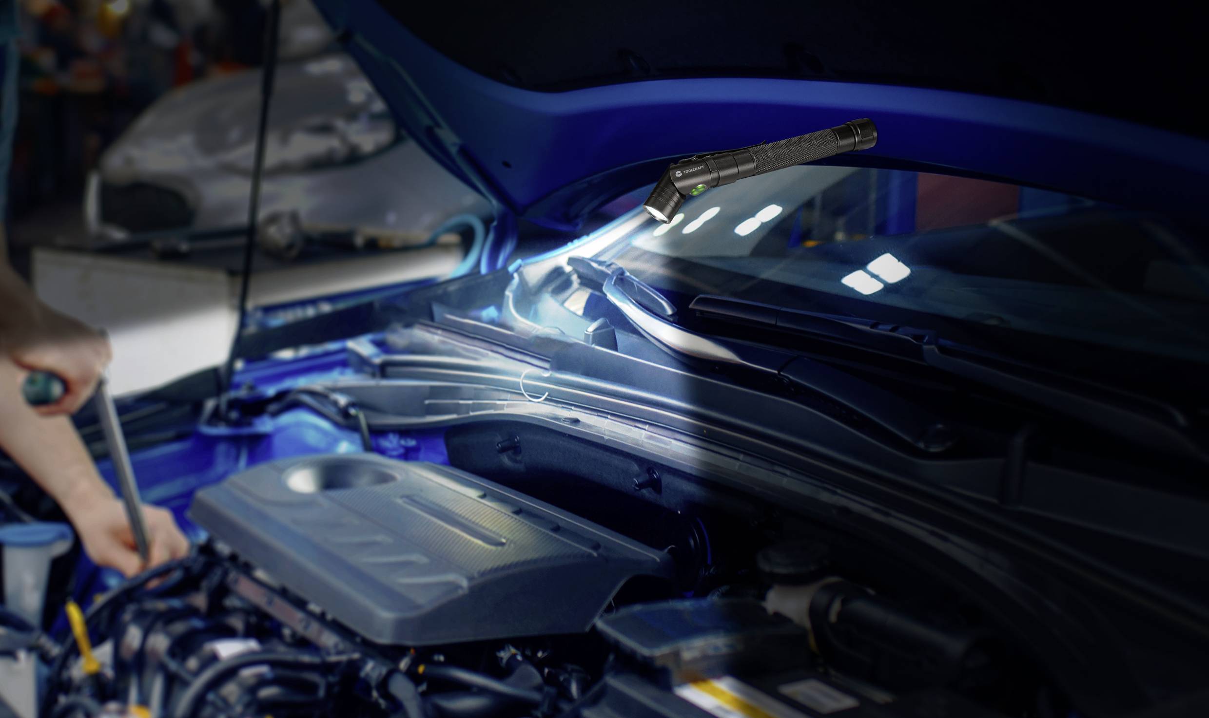 Ein Mechaniker arbeitet unter der geöffneten Motorhaube eines Autos, beleuchtet von einer Lampe. Der Fokus liegt auf technischen Details.