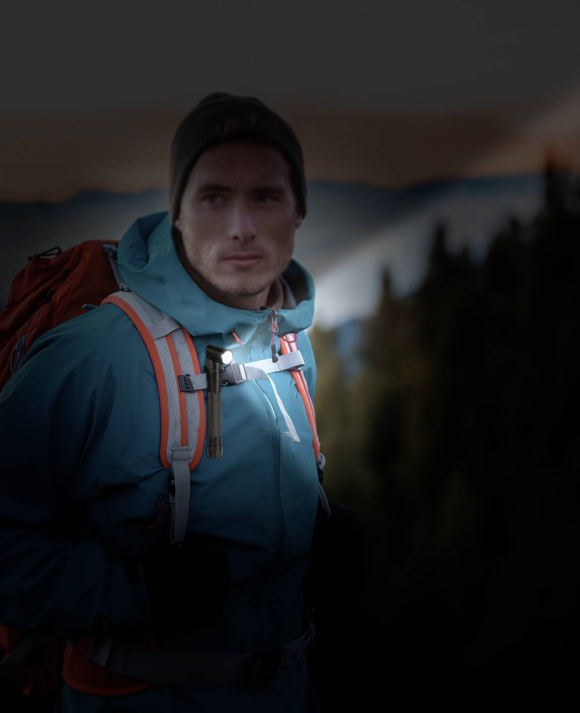 Ein Mann in Outdoor-Kleidung mit Rucksack steht in einer bergigen Landschaft bei Dämmerung. Er blickt aufmerksam in die Ferne.