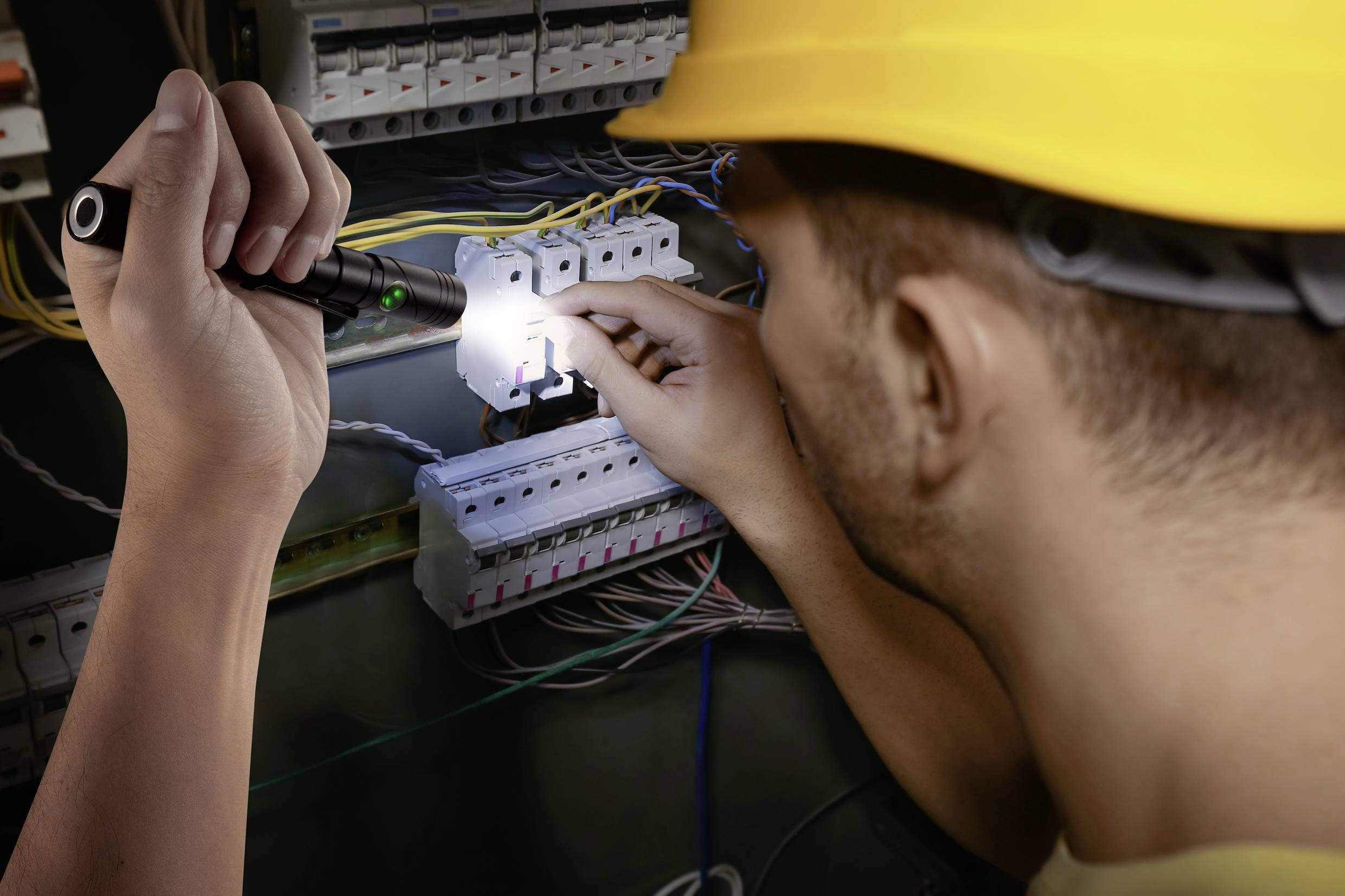 Ein Elektriker mit Schutzhelm arbeitet an einem geöffneten Schaltschrank. Er hält eine Taschenlampe, die den Sicherungskasten beleuchtet.
