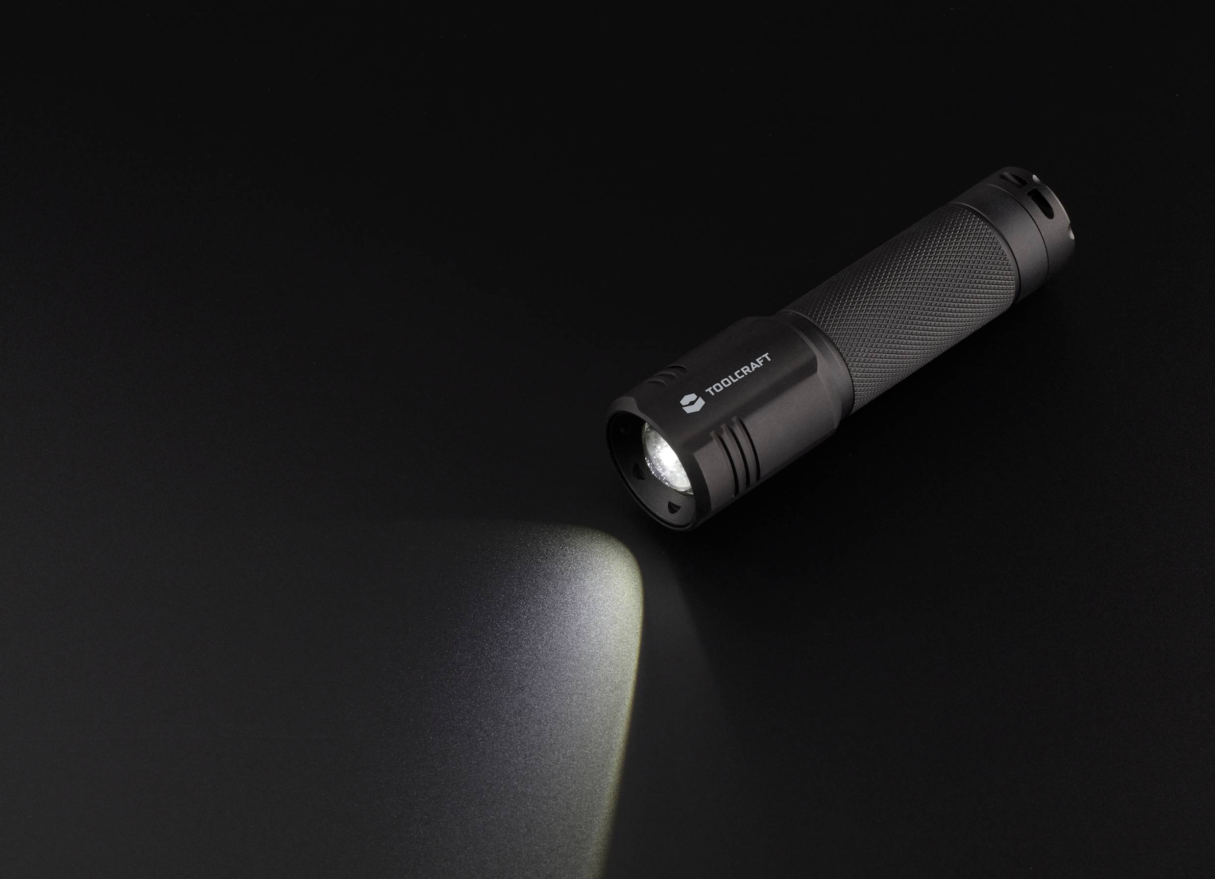 Taschenlampe auf schwarzem Hintergrund leuchtet schräg nach unten. Die Lichtstrahlung wirft einen hellen Kegel auf die dunkle Fläche.