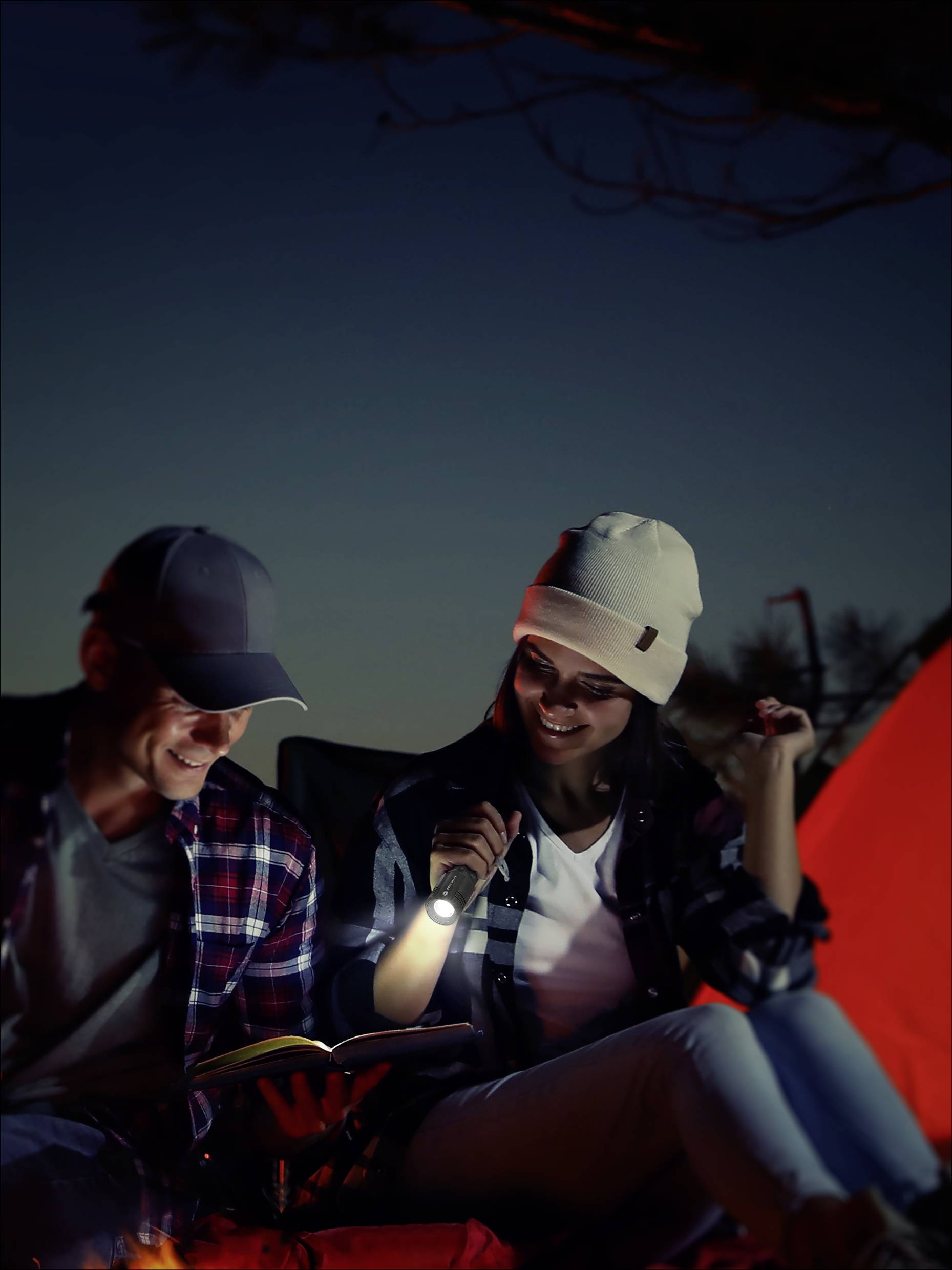 Ein Mann und eine Frau, die gemütlich beim Camping im Freien sitzen. Sie lachen und lesen bei Laternenlicht vor einem Zelt.