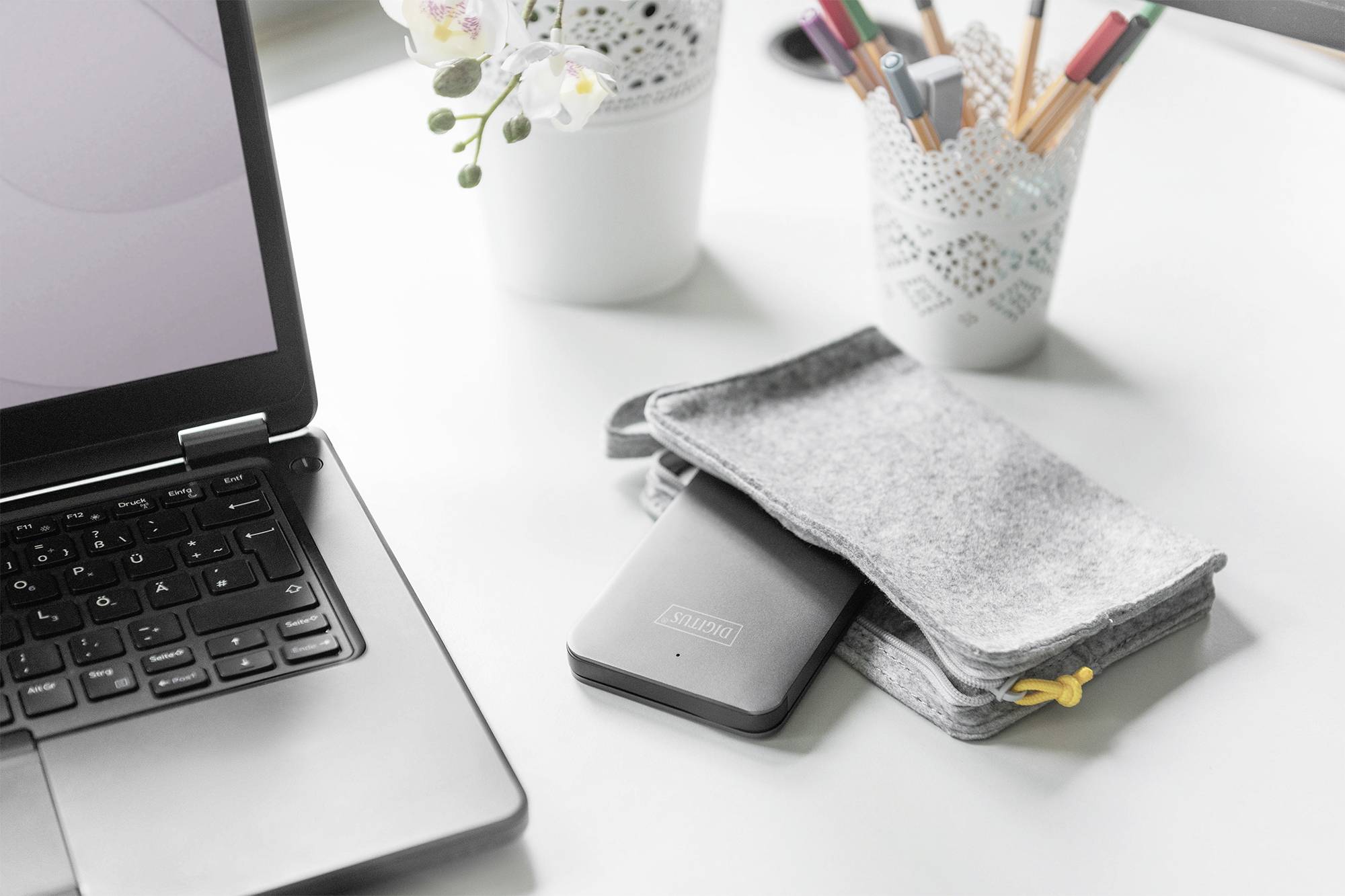 Ein Laptop steht offen auf einem weißen Tisch. Daneben liegen ein graues Filztäschchen und externe Festplatte. Im Hintergrund Stifte und eine Pflanze.