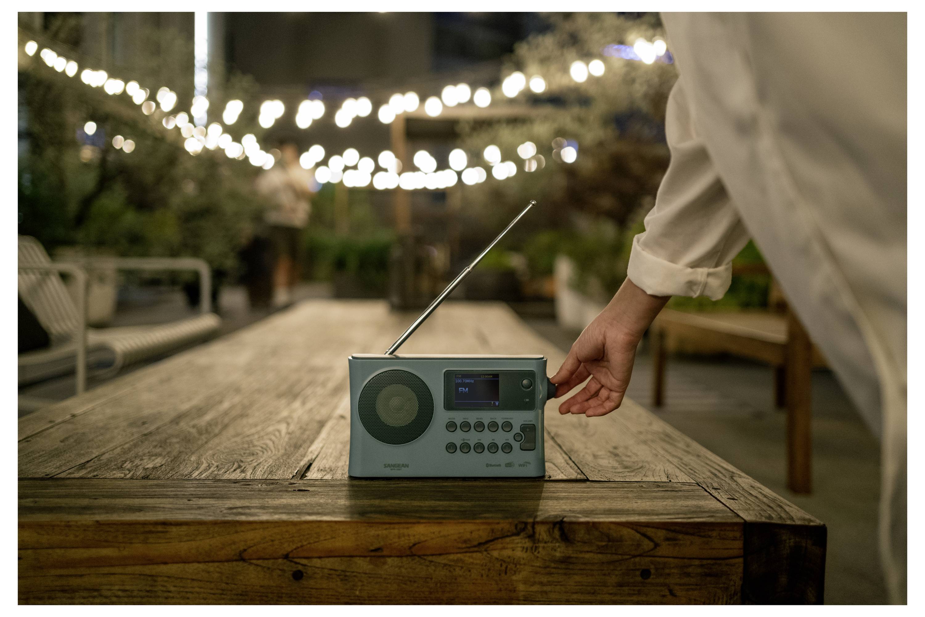 Eine Hand legt ein türkises tragbares Radio auf einen Holztisch im Freien unter Lichterketten in der Dämmerung.