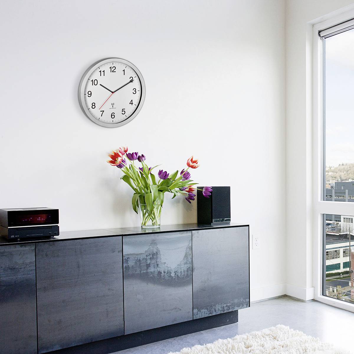 Eine moderne Inneneinrichtung mit einer schwarzen Kommode, darauf ein Blumenstrauß in einer Vase, eine rote Uhr an der Wand.