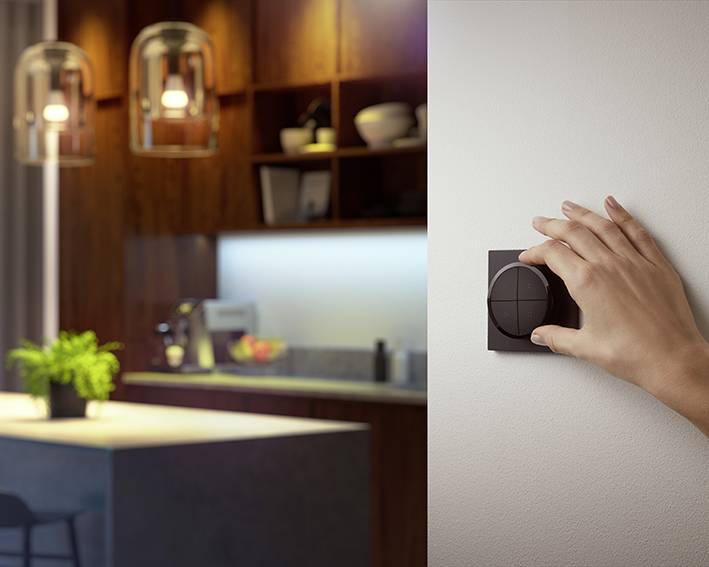 Eine Hand dreht einen Lichtdimmer an der Wand. Im Hintergrund eine modern beleuchtete Küche mit Pflanzen und Küchengeräten.