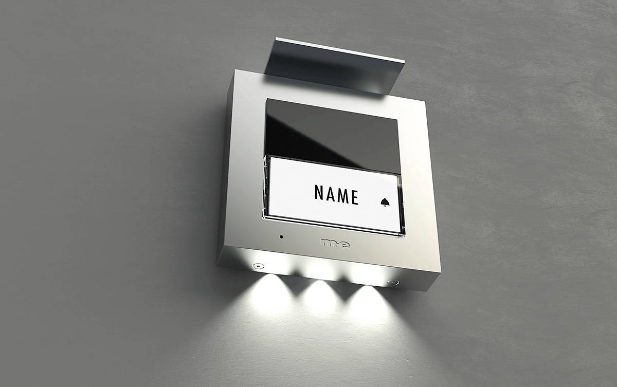 Metallene Türsprechanlage mit Leuchtschild und dem Text 'NAME', darunter drei LED-Leuchten zur Beleuchtung, an einer grauen Wand.