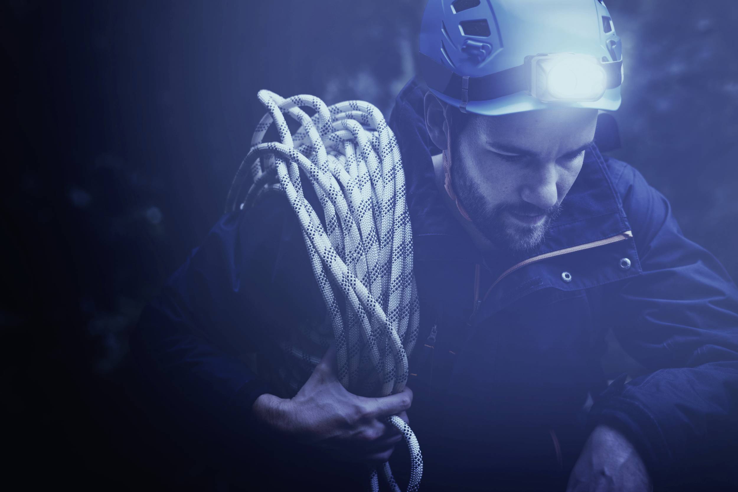Ein Mann mit Kletterausrüstung und Stirnlampe hält ein Seil in einem dunklen, unbekannten Umfeld.