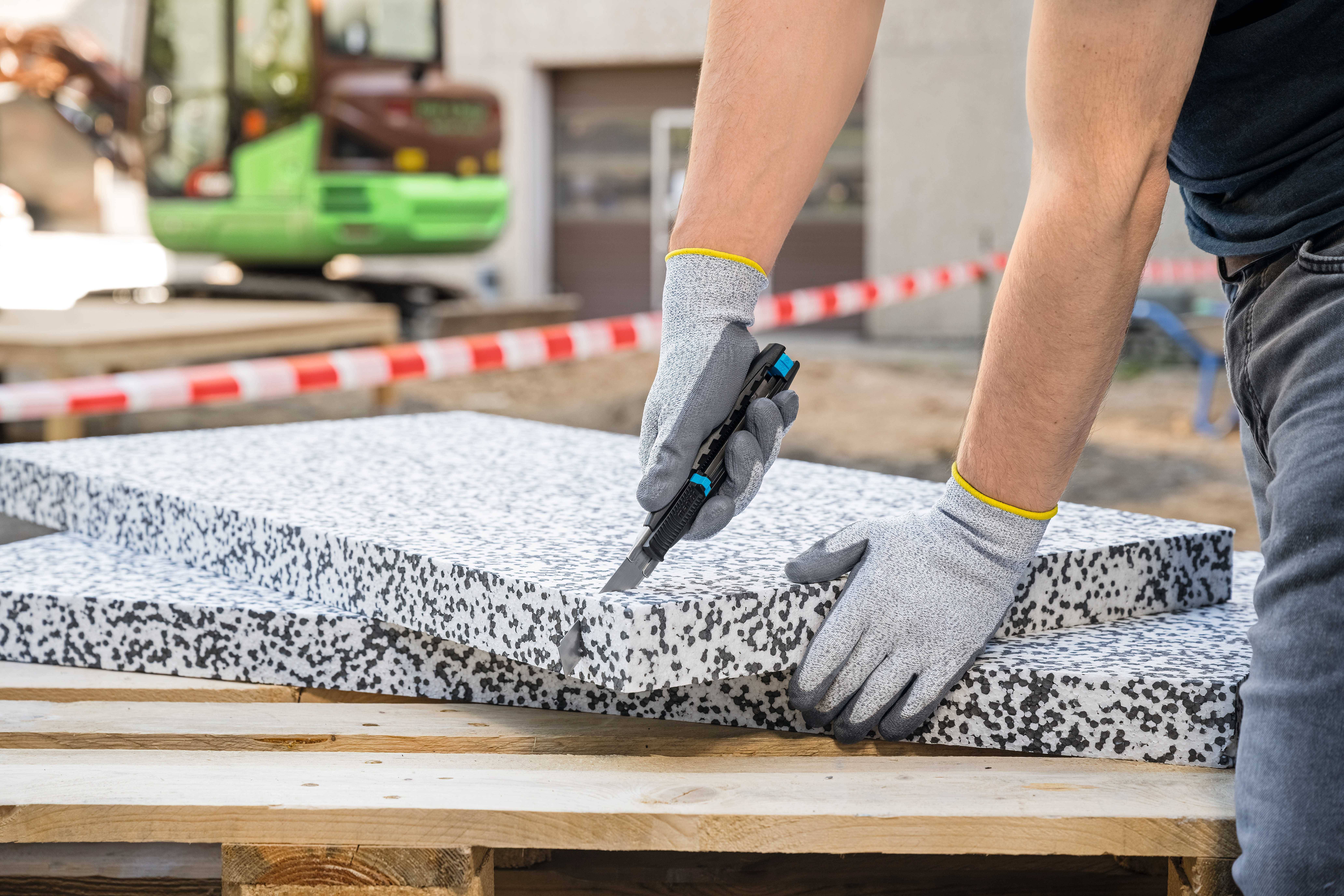 Eine Person in Arbeitshandschuhen schneidet mit einem Cutter-Messer eine Isolierplatte auf einer Baustelle.