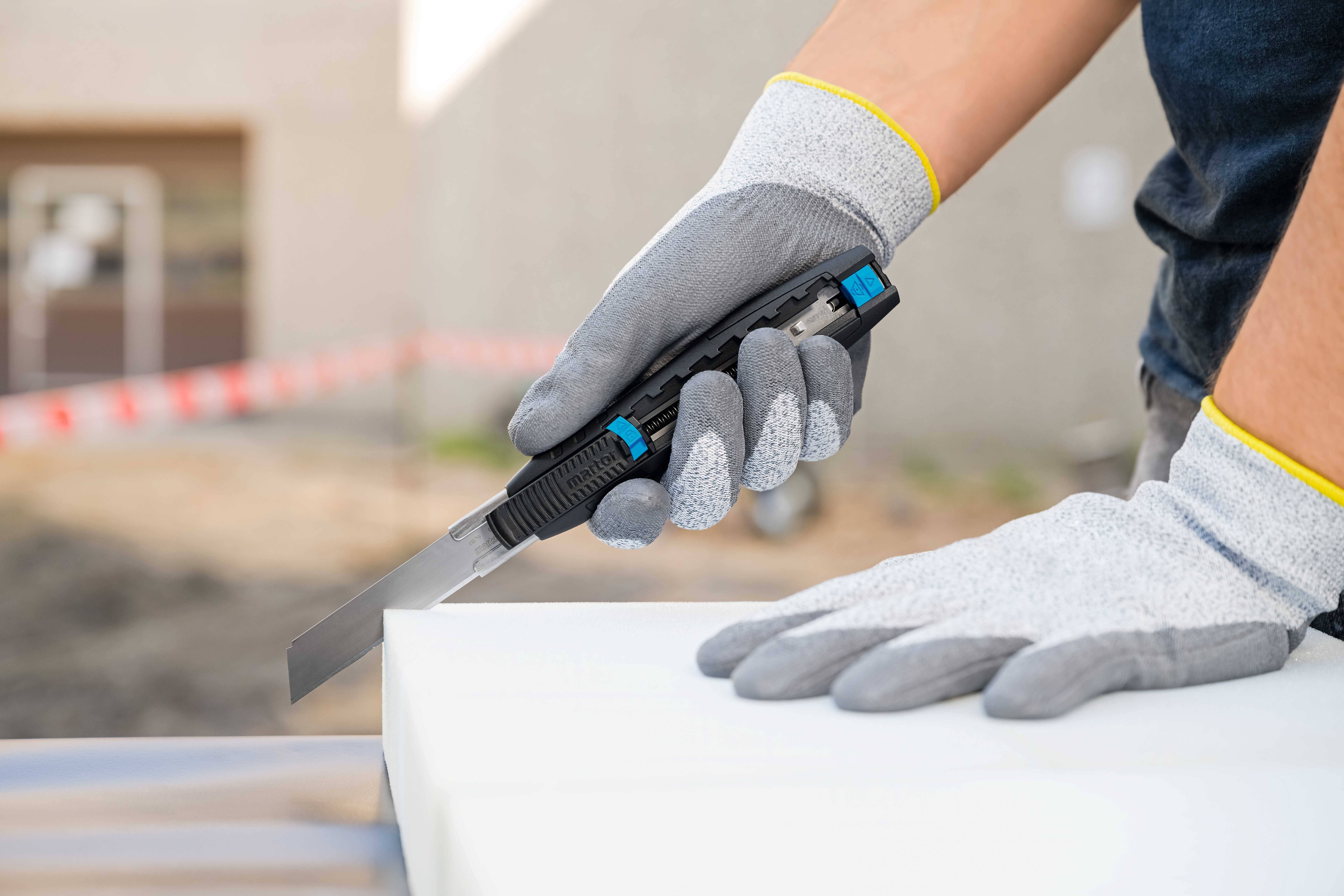 Person schneidet Material mit einem Cuttermesser, trägt graue Handschuhe. Thema: Heimwerken oder Handwerksarbeit.