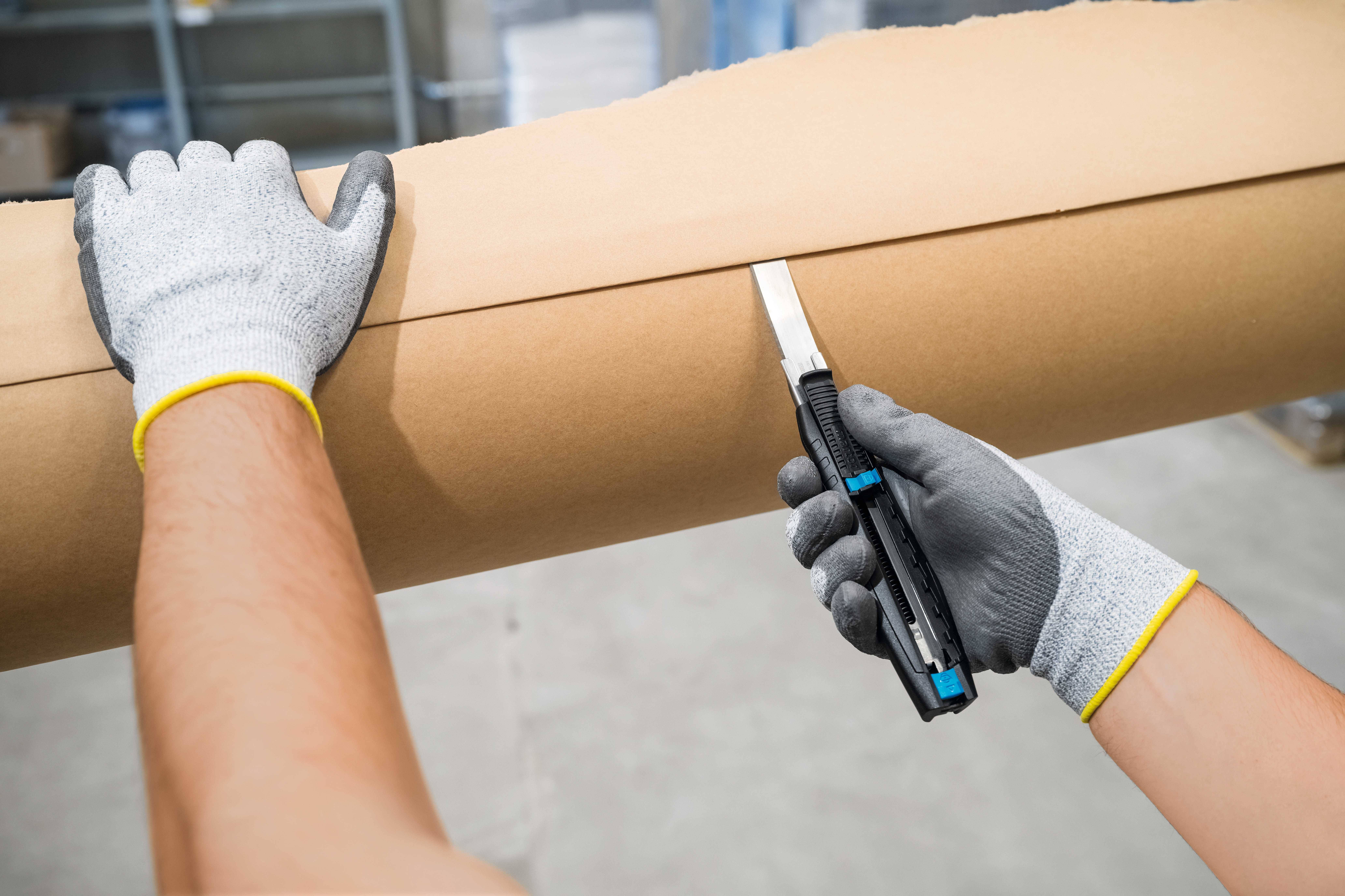 Eine Person mit grauen Arbeitshandschuhen schneidet mit einem Cutter-Messer braunes Verpackungspapier auf einer Rolle.