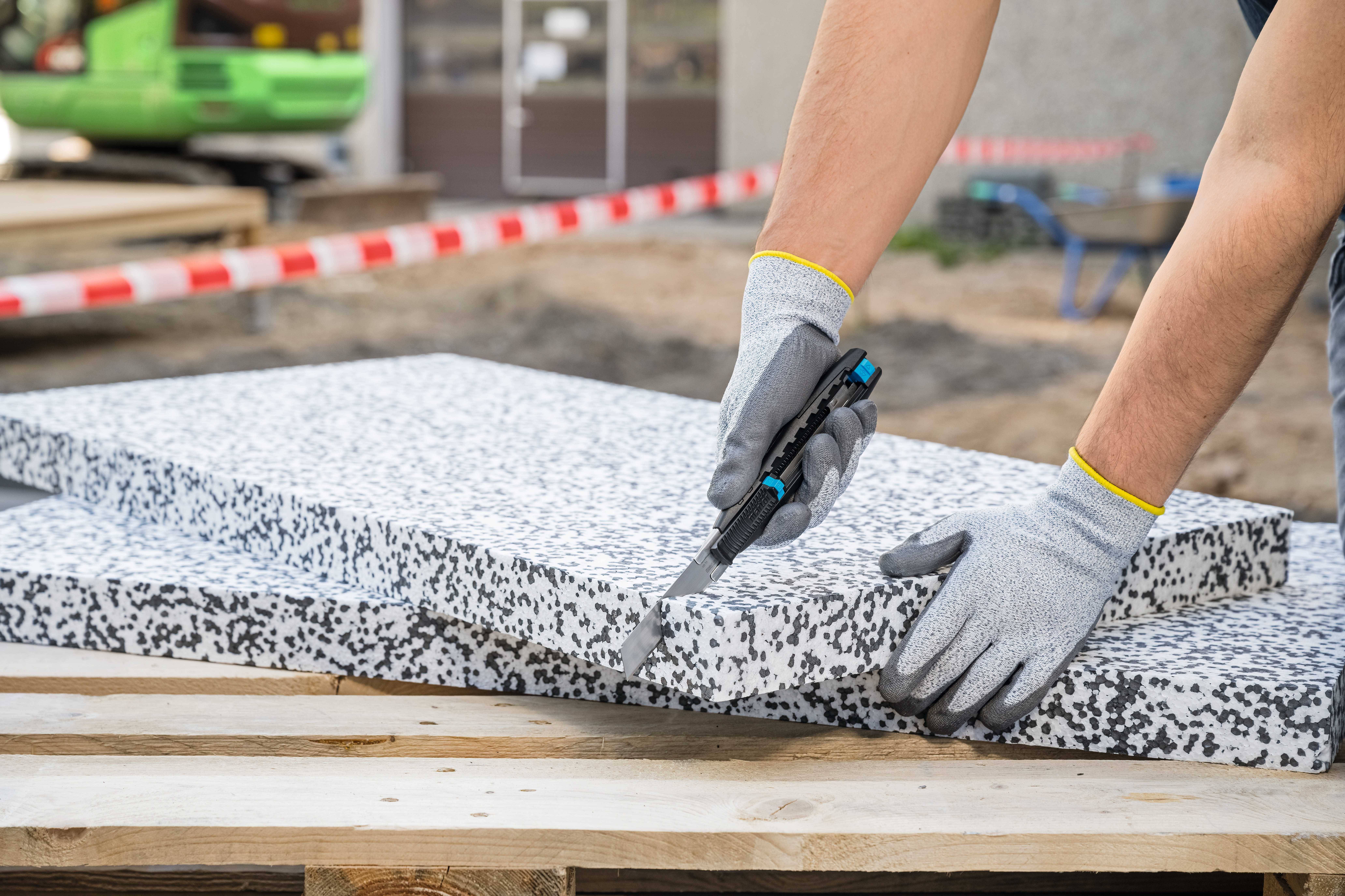 Eine Person schneidet eine Dämmplatte aus Polystyrol mit einem Messer auf einer Baustelle. Im Hintergrund sind Baugeräte zu sehen.