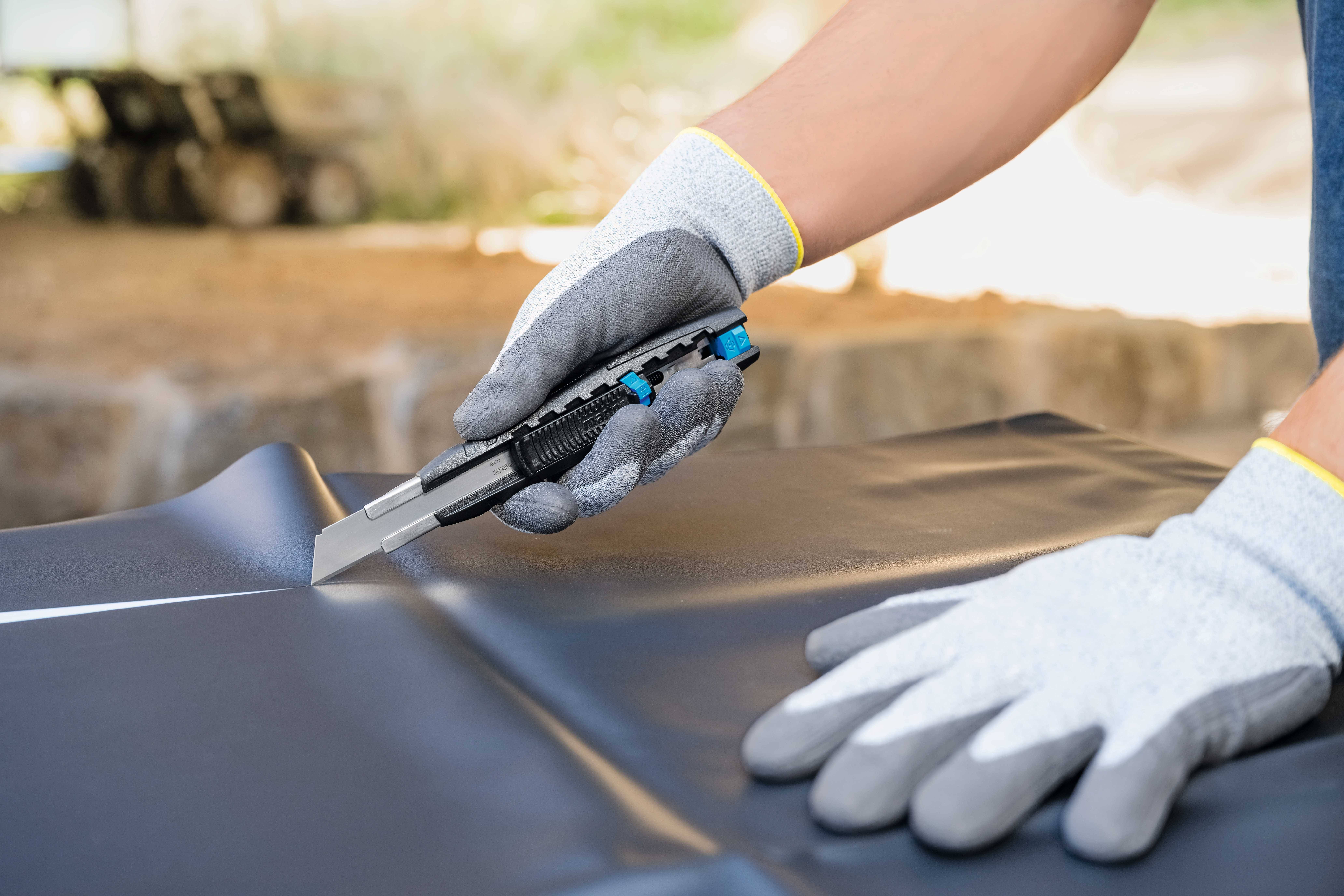 Eine Person mit grauen Arbeitshandschuhen schneidet mit einem Cutter-Messer ein Stück schwarzes Material auf einem Tisch.