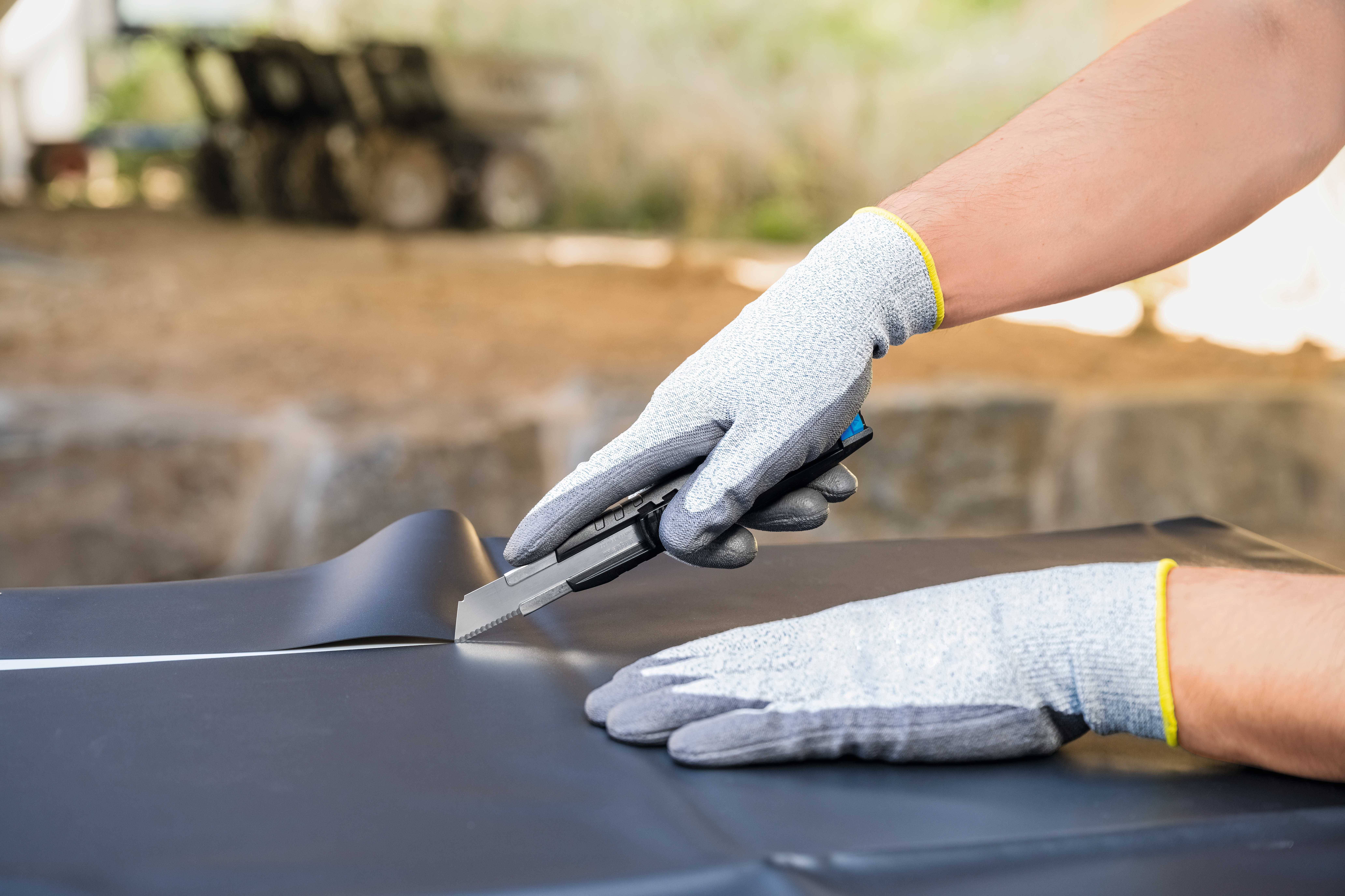 Person mit grauen Arbeitshandschuhen schneidet mit einem Teppichmesser in schwarzes Material. Hintergrund unscharf.