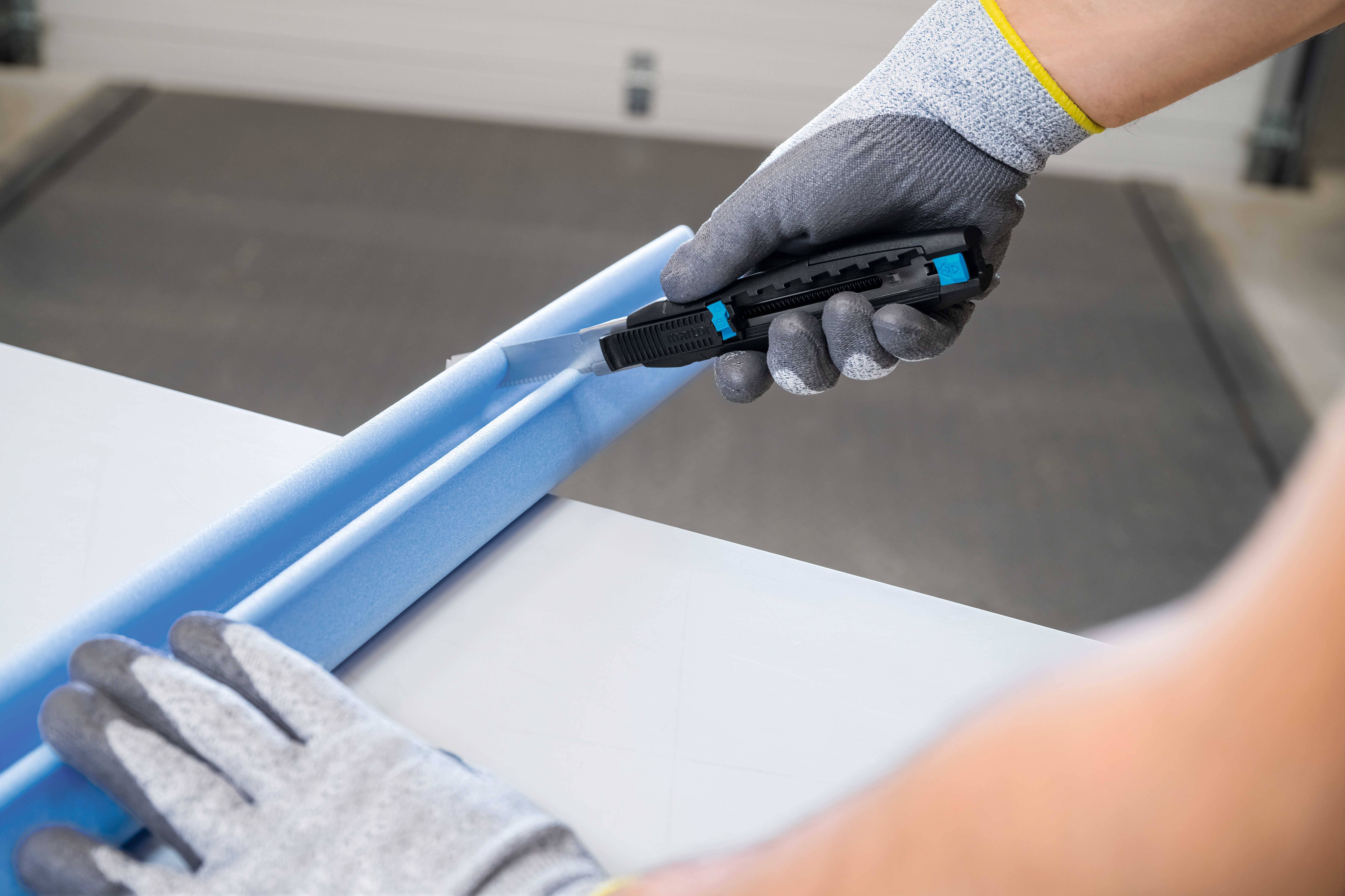 Eine Person schneidet mit einem Cuttermesser blauen Schaumstoff. Sie trägt graue Handschuhe.