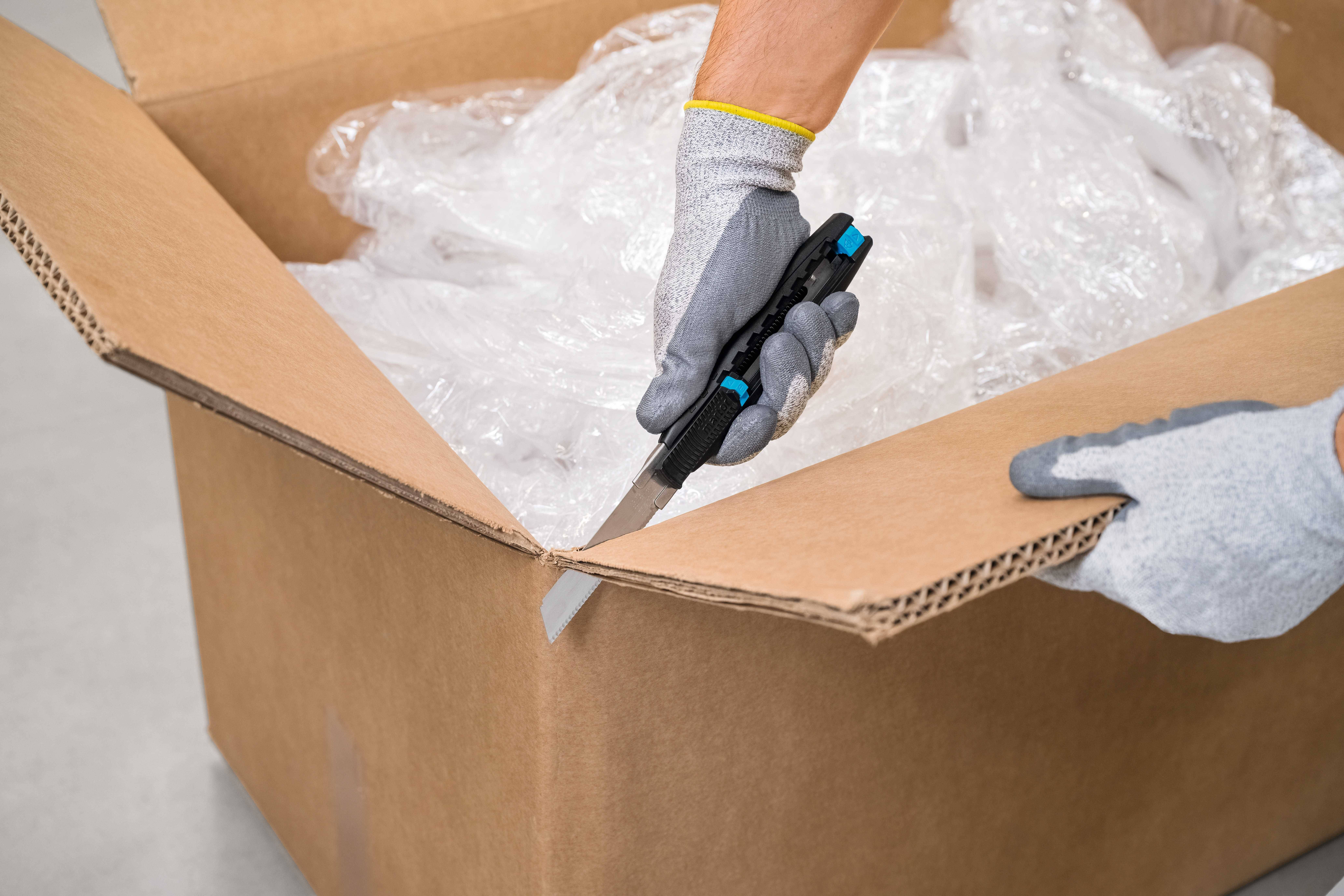 Person öffnet Karton mit Cutter-Messer, innen sind Kunststoffverpackungen.
