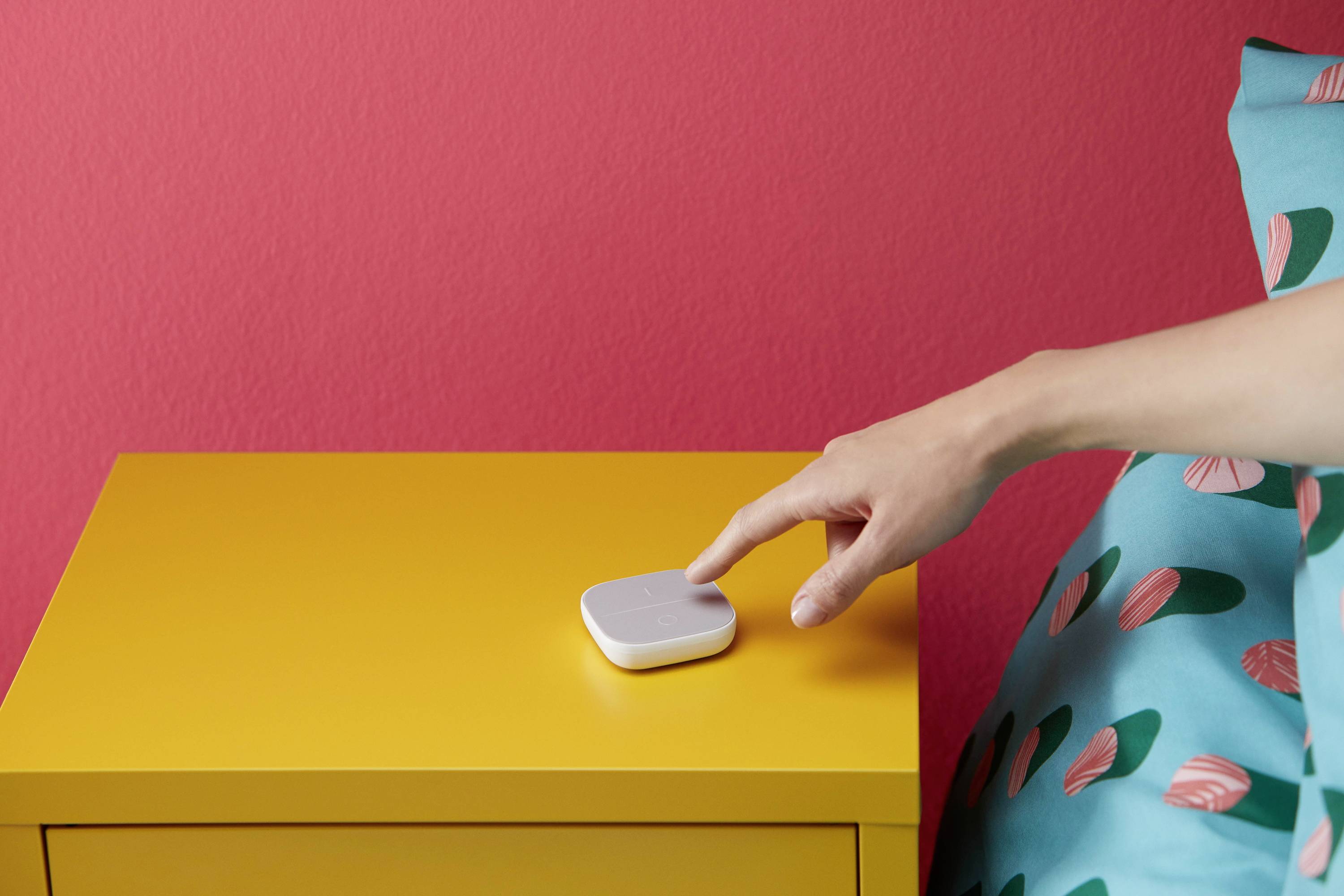 Eine Hand schaltet ein kleines, weißes Gerät auf einem gelben Nachttisch ein. Der Hintergrund ist eine rote Wand und ein buntes Bettkissen.