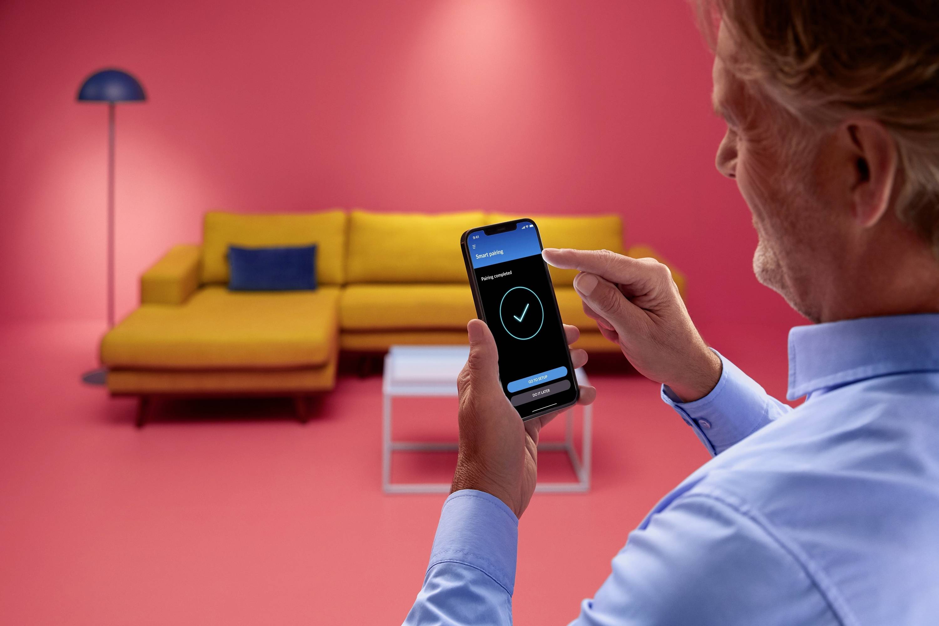 Eine Person bedient ein Smartphone mit einer App in einem Raum mit gelbem Sofa und rosa Wänden. Ein weißer Tisch und eine Lampe sind sichtbar.