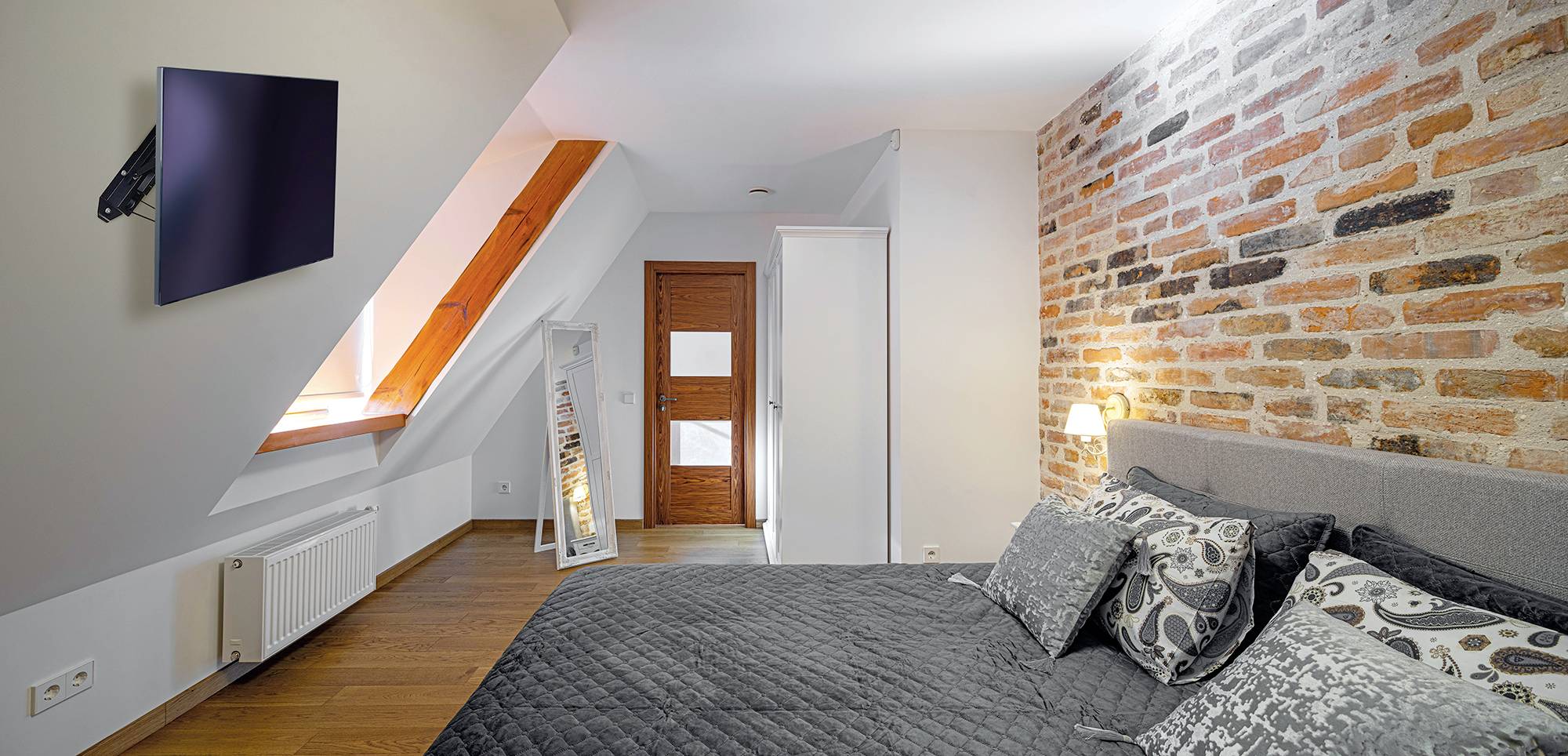 Gemütliches Schlafzimmer mit Backsteinwand, modernem Flachbildfernseher an der Wand, grauem Doppelbett mit gemusterten Kissen.