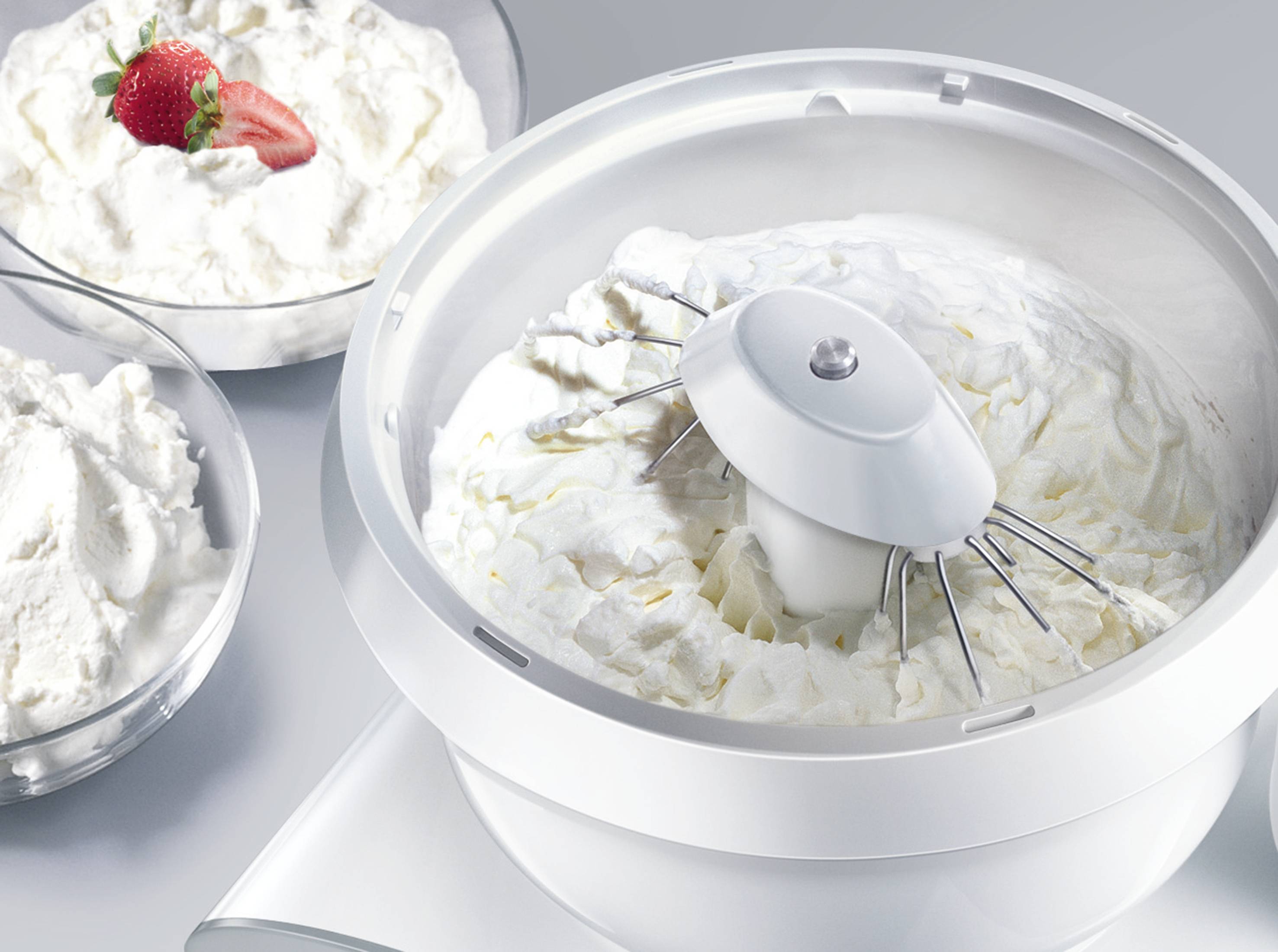 Eine Küchenmaschine schlägt Sahne steif. Im Hintergrund stehen Schüsseln mit fertiger Schlagsahne, eine mit einer Erdbeergarnitur.