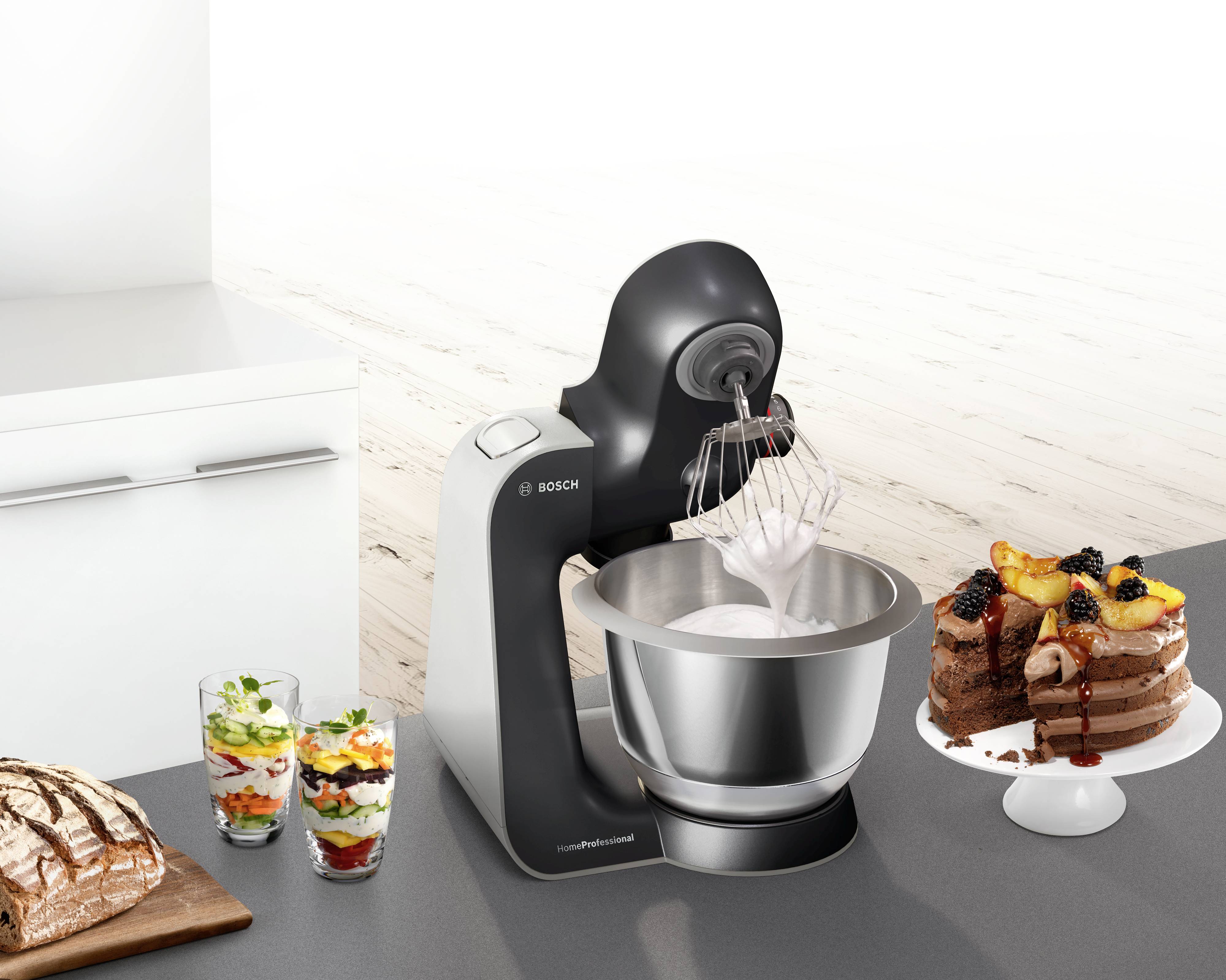 Eine Küchenmaschine rührt eine Masse in einer Metallschüssel. Daneben ein Kuchen, zwei Dessertgläser und ein Laib Brot auf der Arbeitsplatte.