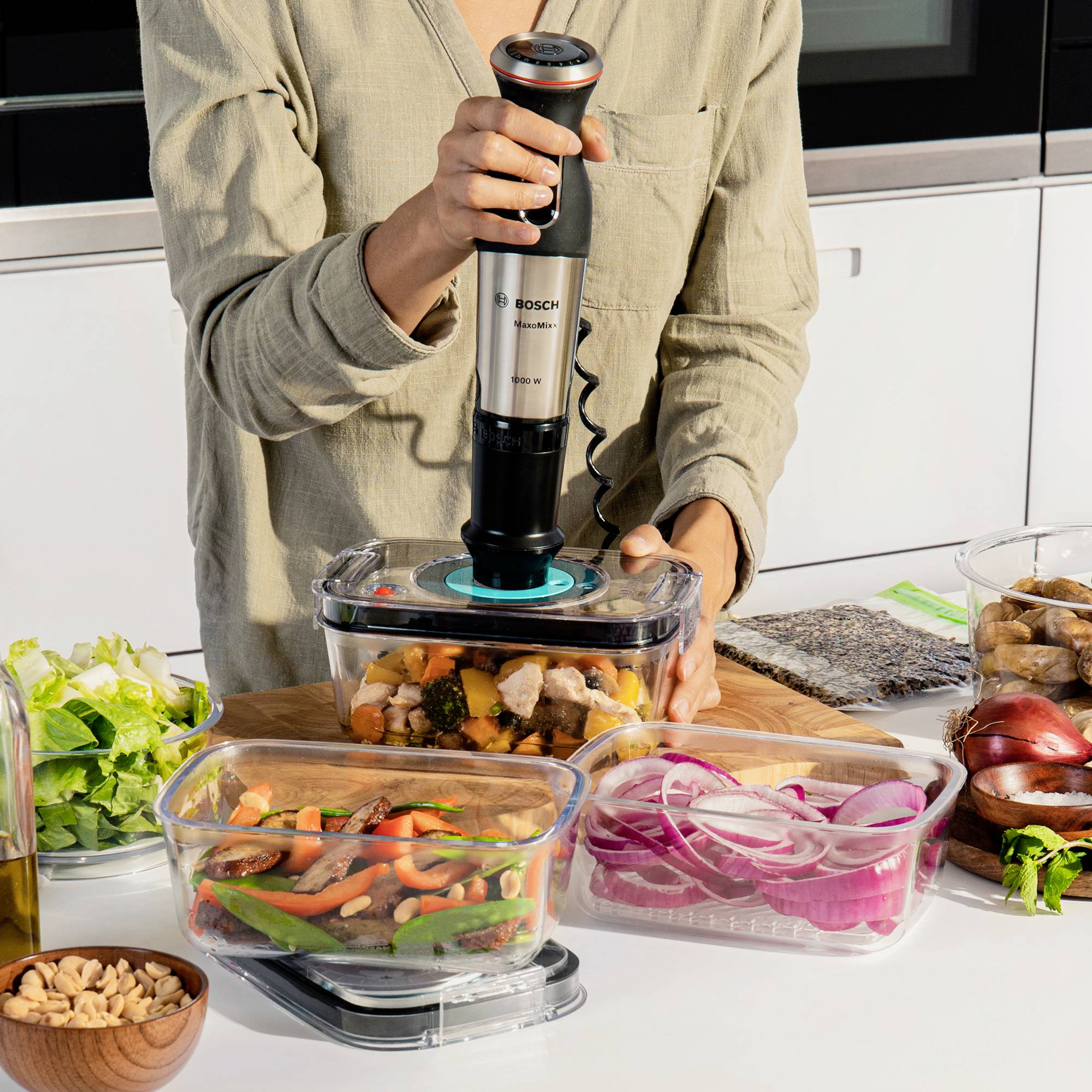Person benutzt einen Stabmixer, um Gemüse in einer Schüssel zu pürieren. Auf dem Tisch liegen verschiedene Lebensmittel und Zutaten.