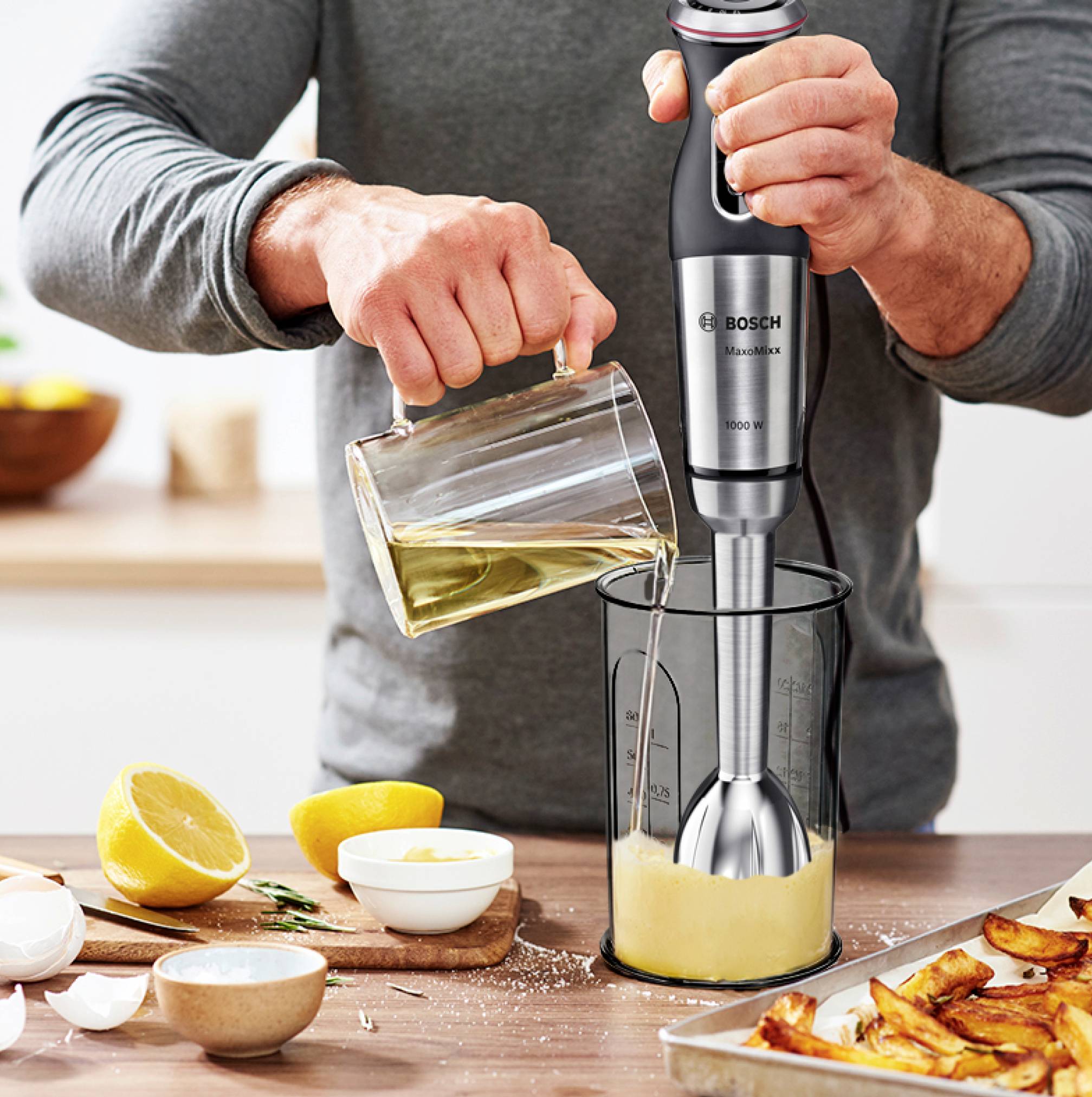 Eine Person benutzt einen Stabmixer, um Flüssigkeit in einem Behälter zu pürieren. Daneben liegen Zitronen, Eier und eine Backform.