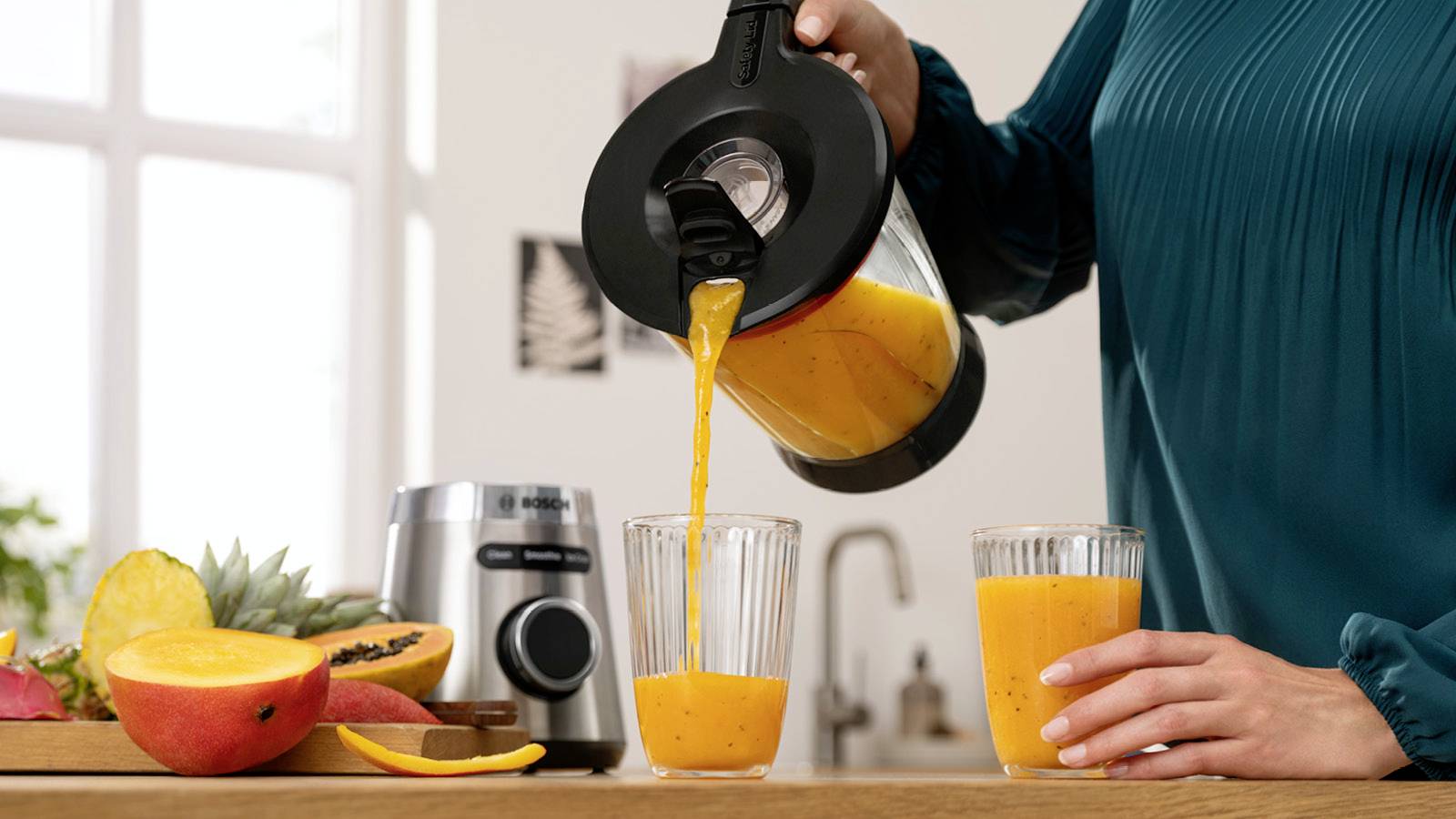 Eine Person gießt frischen Orangensaft aus einem Mixer in zwei Gläser. Im Hintergrund sind verschiedene Obstsorten zu sehen.