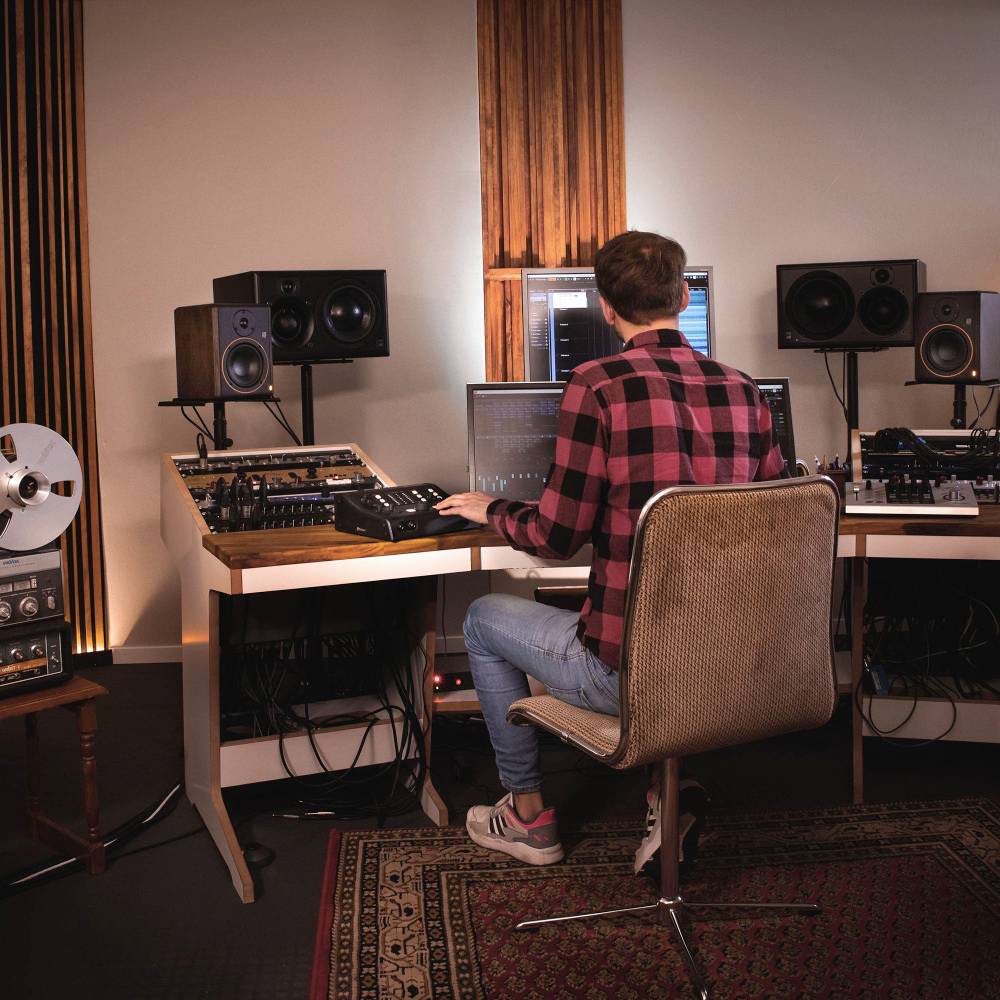 Person in kariertem Hemd arbeitet in einem Studio mit mehreren Monitoren und Lautsprechern; Umgebung deutet auf Musikproduktion hin.