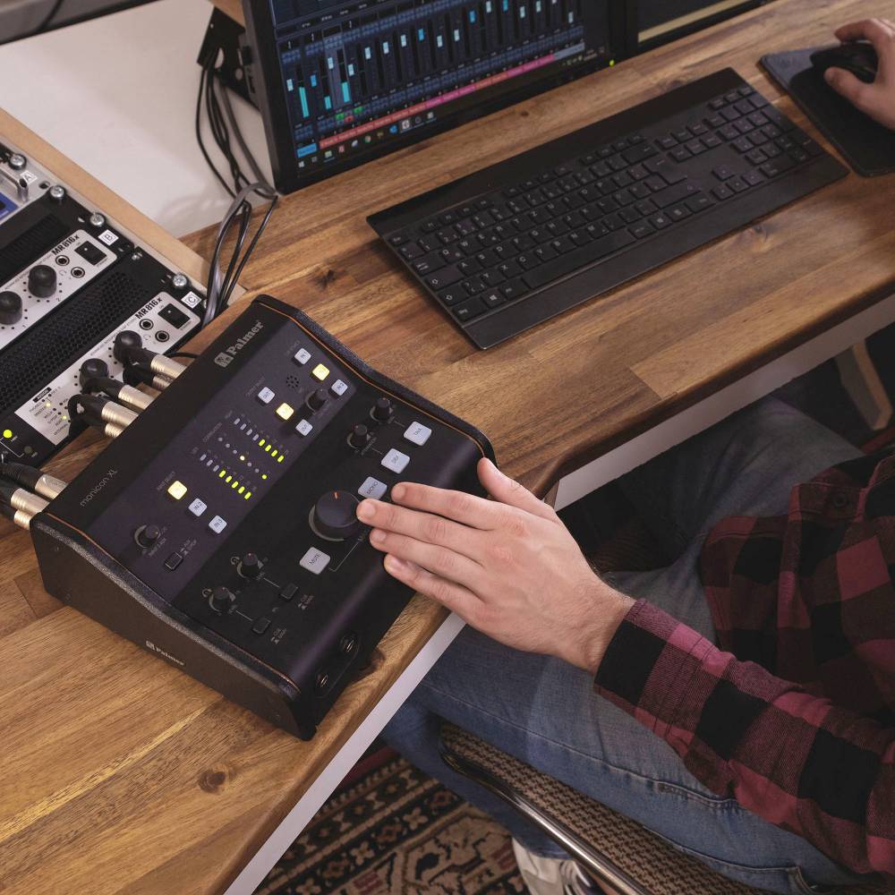 Person an einem Schreibtisch verwendet ein Audiomischpult mit mehreren Reglern und Knöpfen neben Computerbildschirmen und Tastatur.