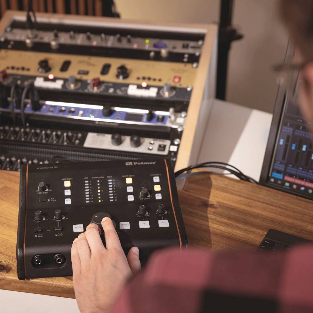 Eine Person bedient ein Audio-Mischpult in einem Studio, umgeben von diversen Audiogeräten und einem Computerbildschirm.