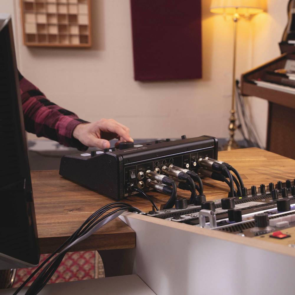 Tonstudio-Aufnahme: Person justiert Audiogeräte an einem Tisch, umgeben von Mischpult und Kabeln. Im Hintergrund eine Lampe und Wanddekoration.