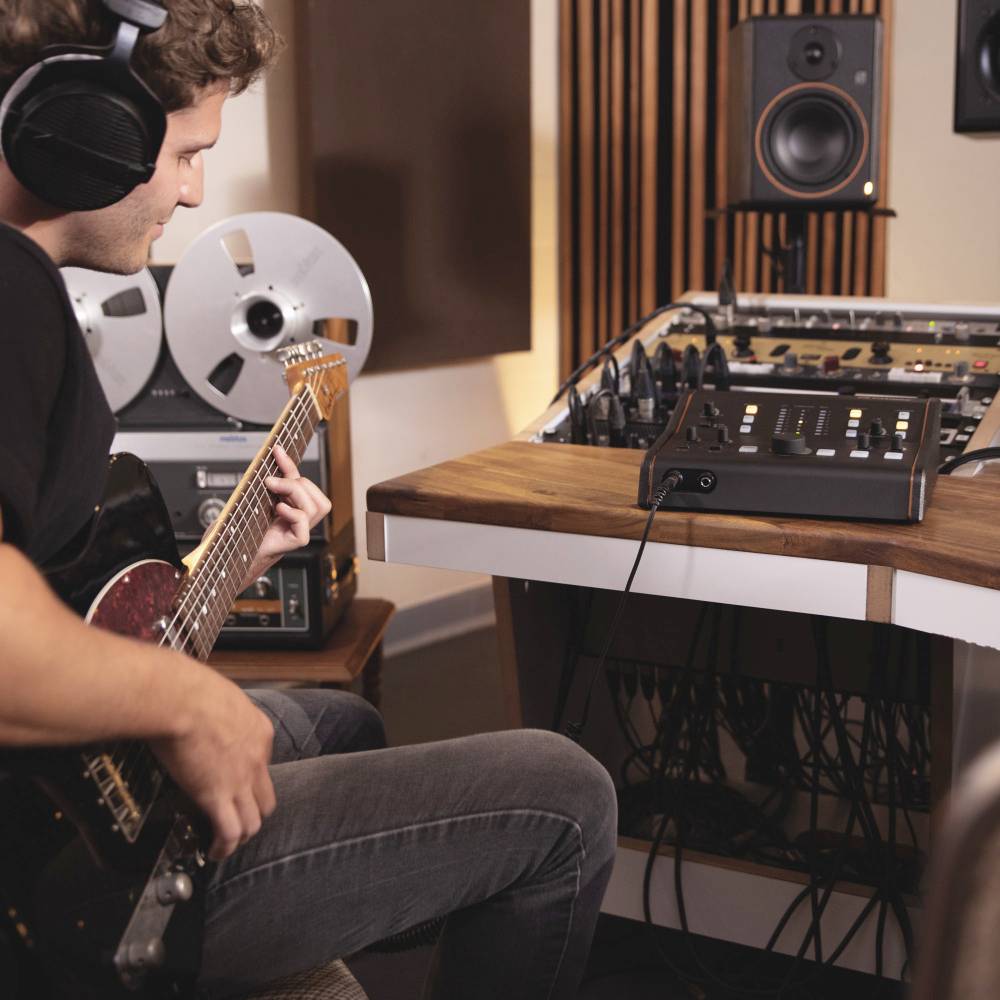 Musiker im Aufnahmestudio spielt E-Gitarre, trägt Kopfhörer. Im Hintergrund Tonbandgeräte und Mischpult.