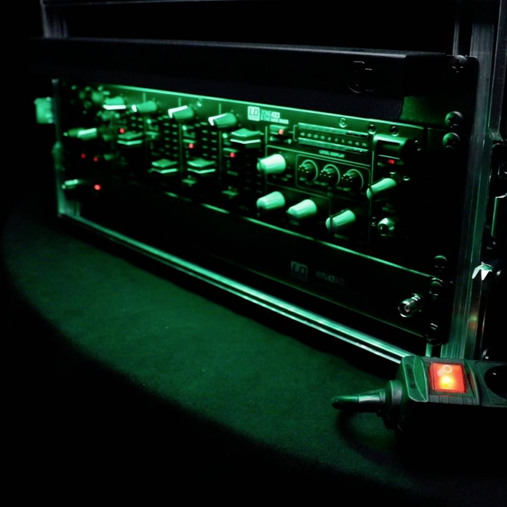 Ein beleuchtetes Audiomischpult in einem dunklen Raum. Pult zeigt mehrere Knöpfe und Regler, die grün und rot leuchten.