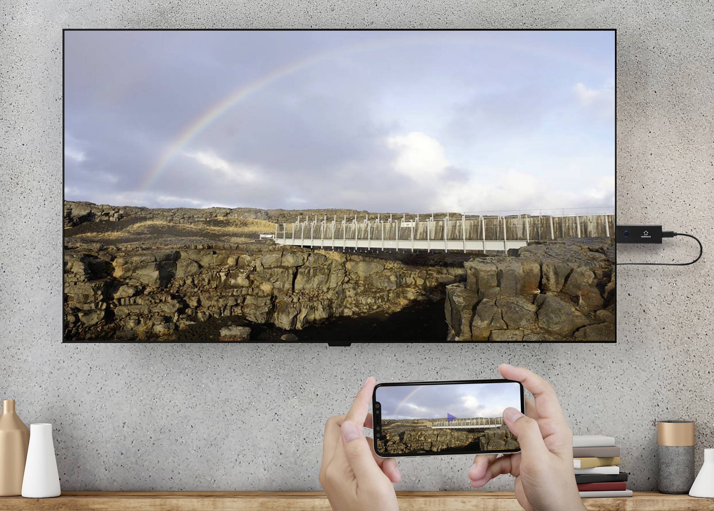 Eine Person streamt ein Naturfoto mit Regenbogen, Felsen und Wolken von einem Smartphone auf einen an der Wand montierten Fernseher.