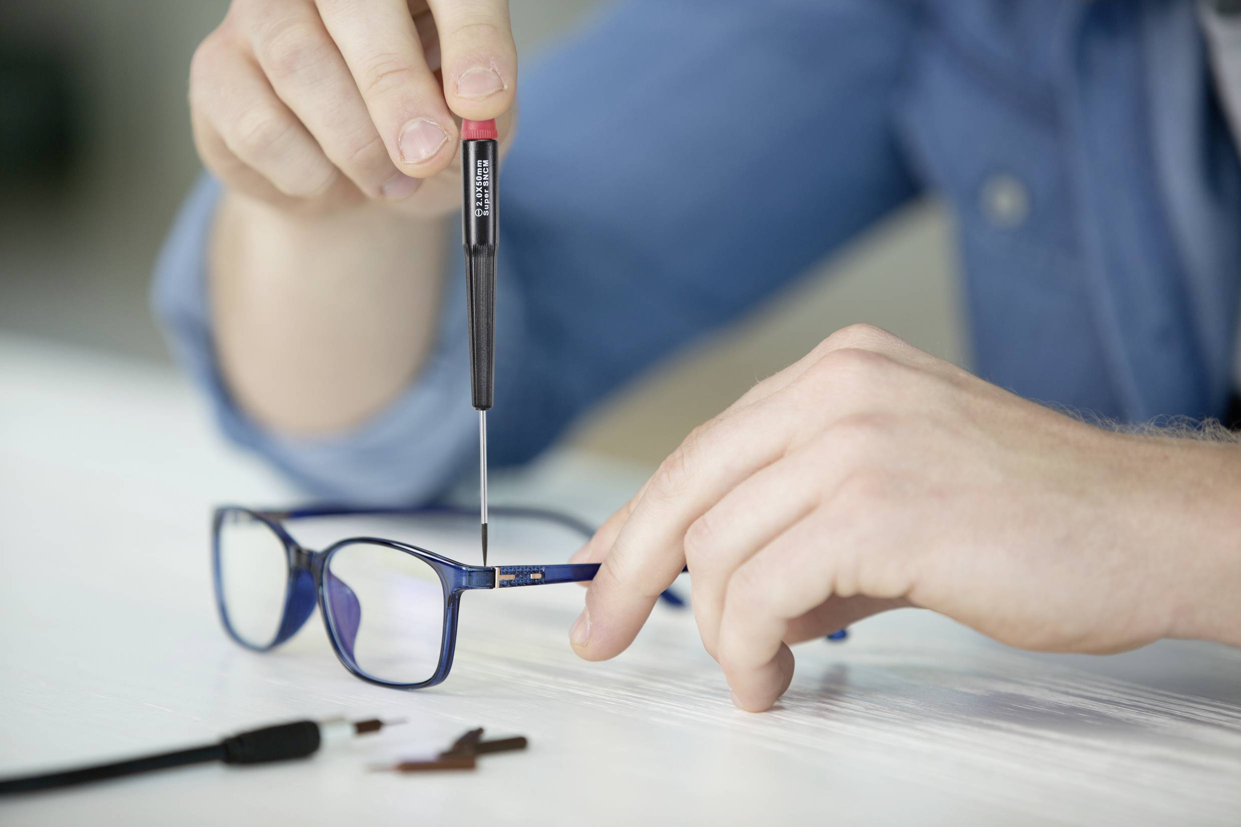 Eine Person repariert eine Brille mit einem kleinen Schraubendreher. Die Brille liegt auf einem Tisch, und Ersatzteile liegen daneben.