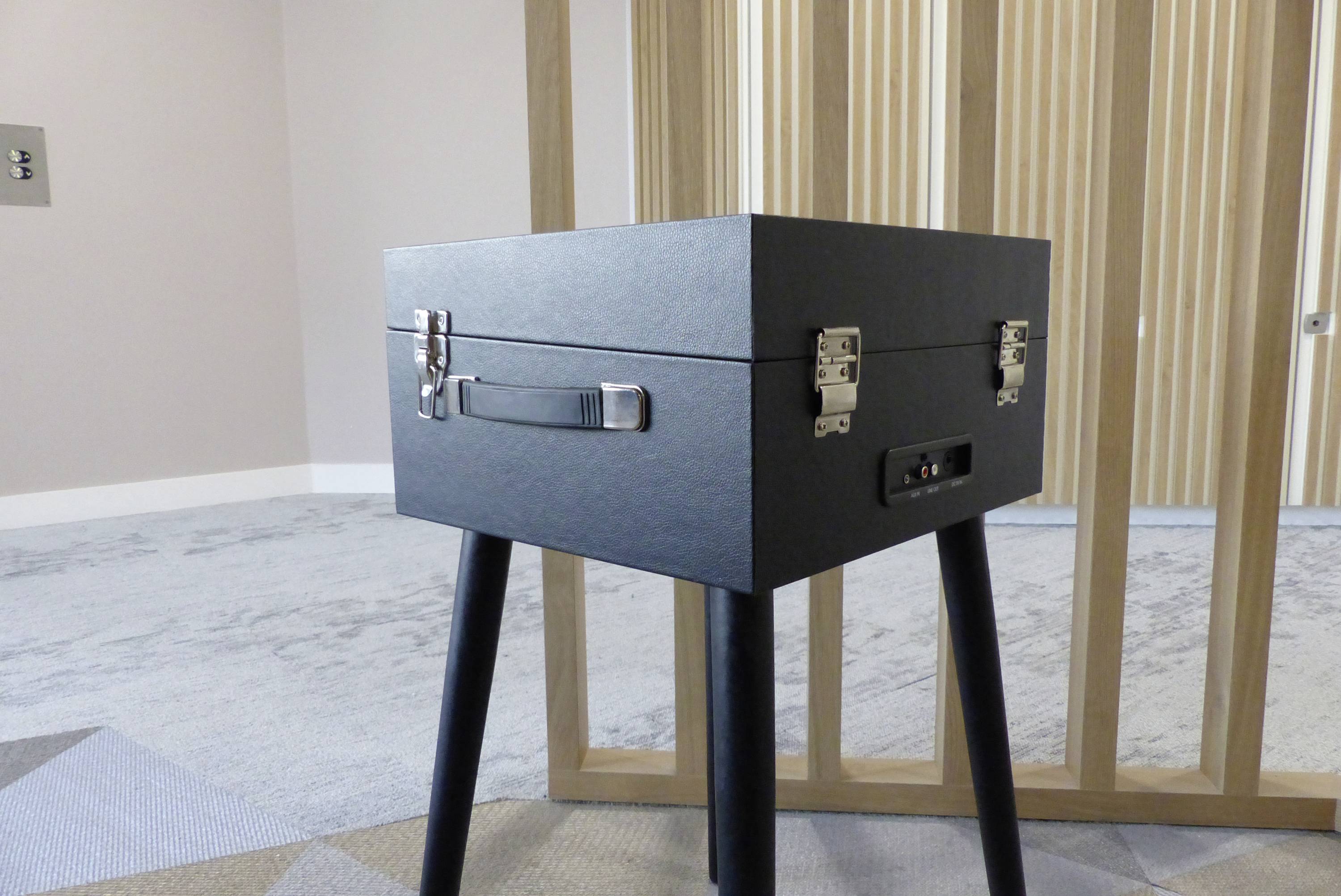 Ein schwarzer Koffer mit Metallverschlüssen steht auf einem schmalen Holztisch, in einem Raum mit teppichartigem Boden und Holzpaneelwänden im Hintergrund.