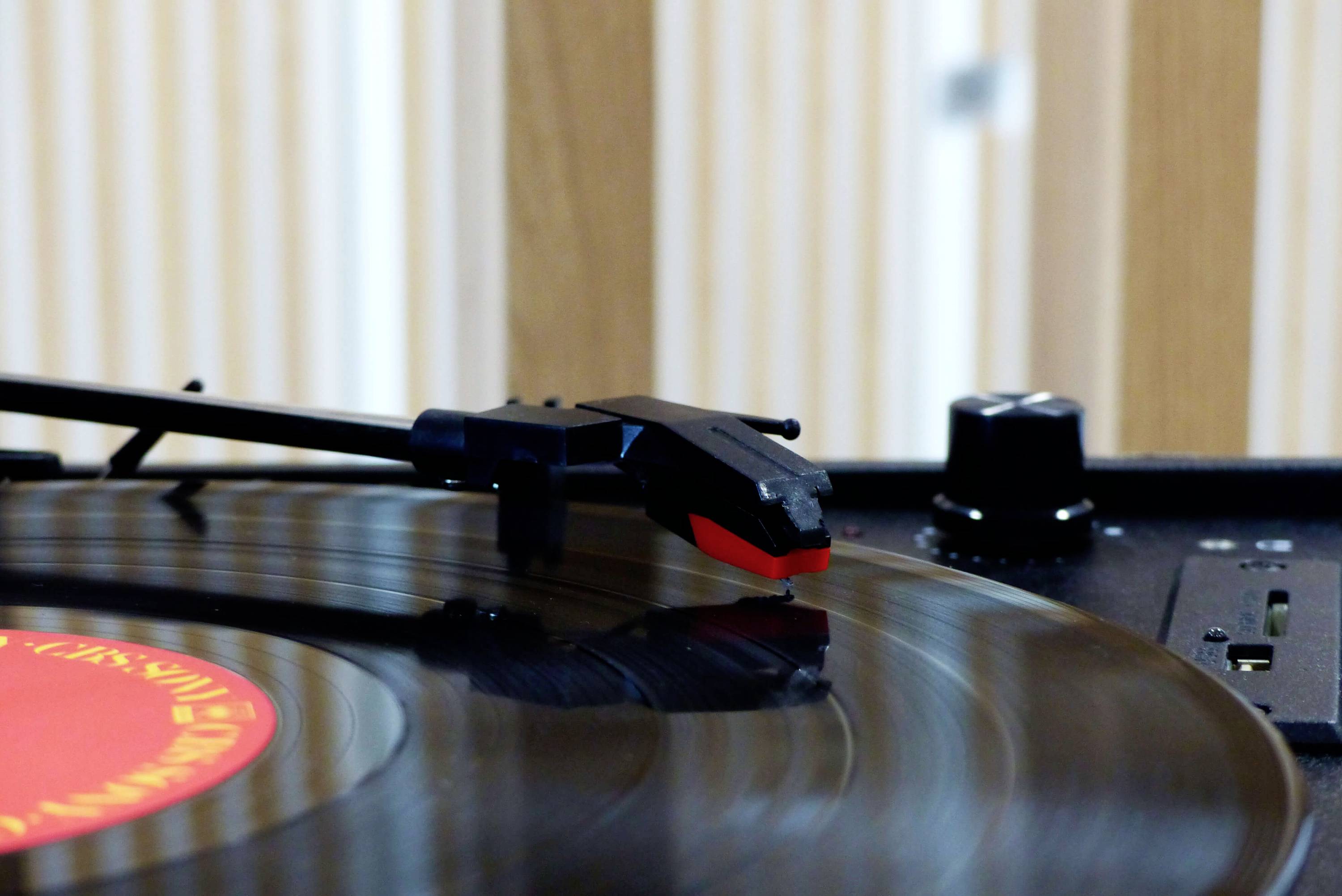 Ein Plattenspieler mit schwarzem Tonarm spielt eine Schallplatte. Im Hintergrund sind unscharfe Holzlatten zu sehen.