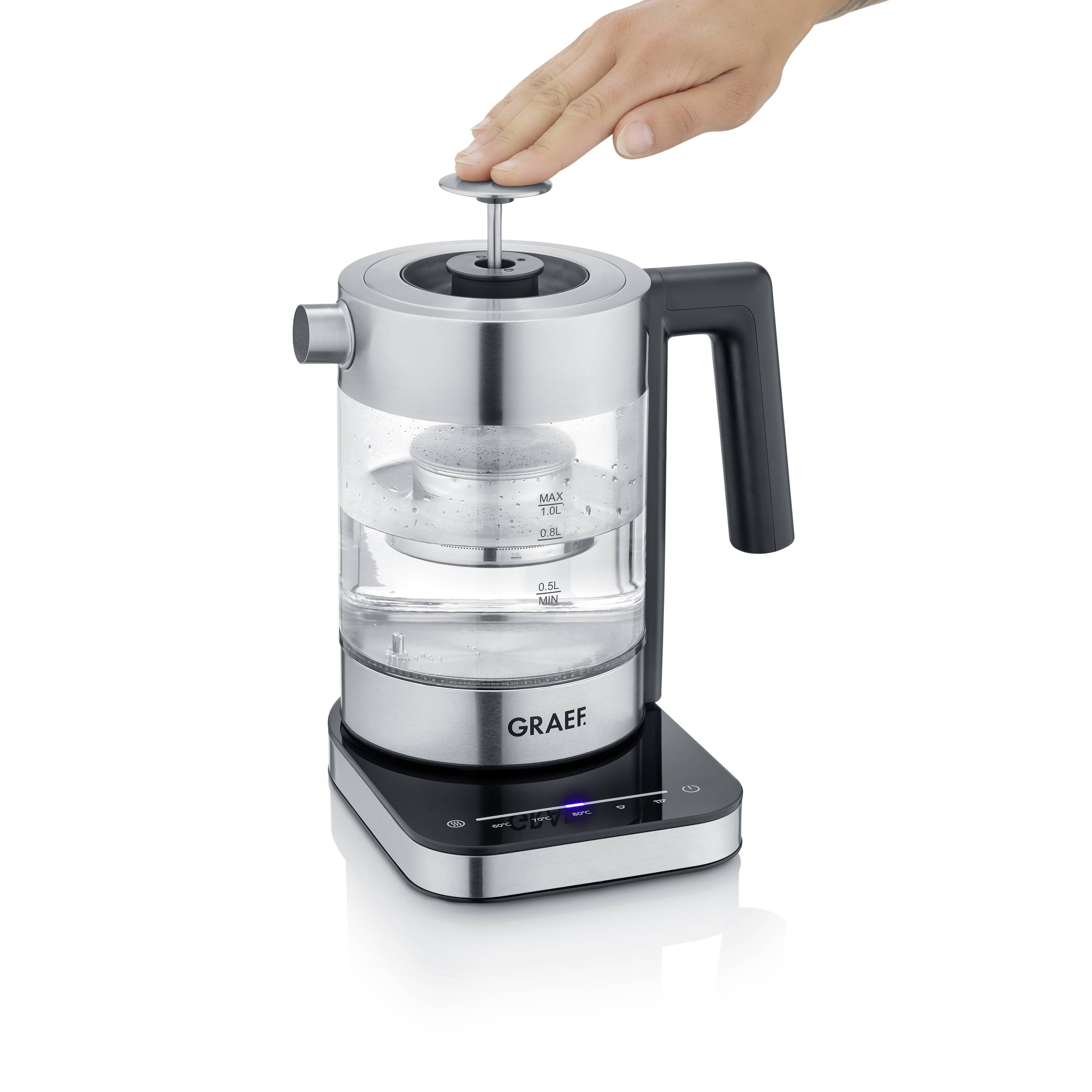 Ein elektrischer Wasserkocher mit Glasbehälter und schwarzem Griff. Eine Hand bedient den Deckel. Marke 'Graef' vorne sichtbar.
