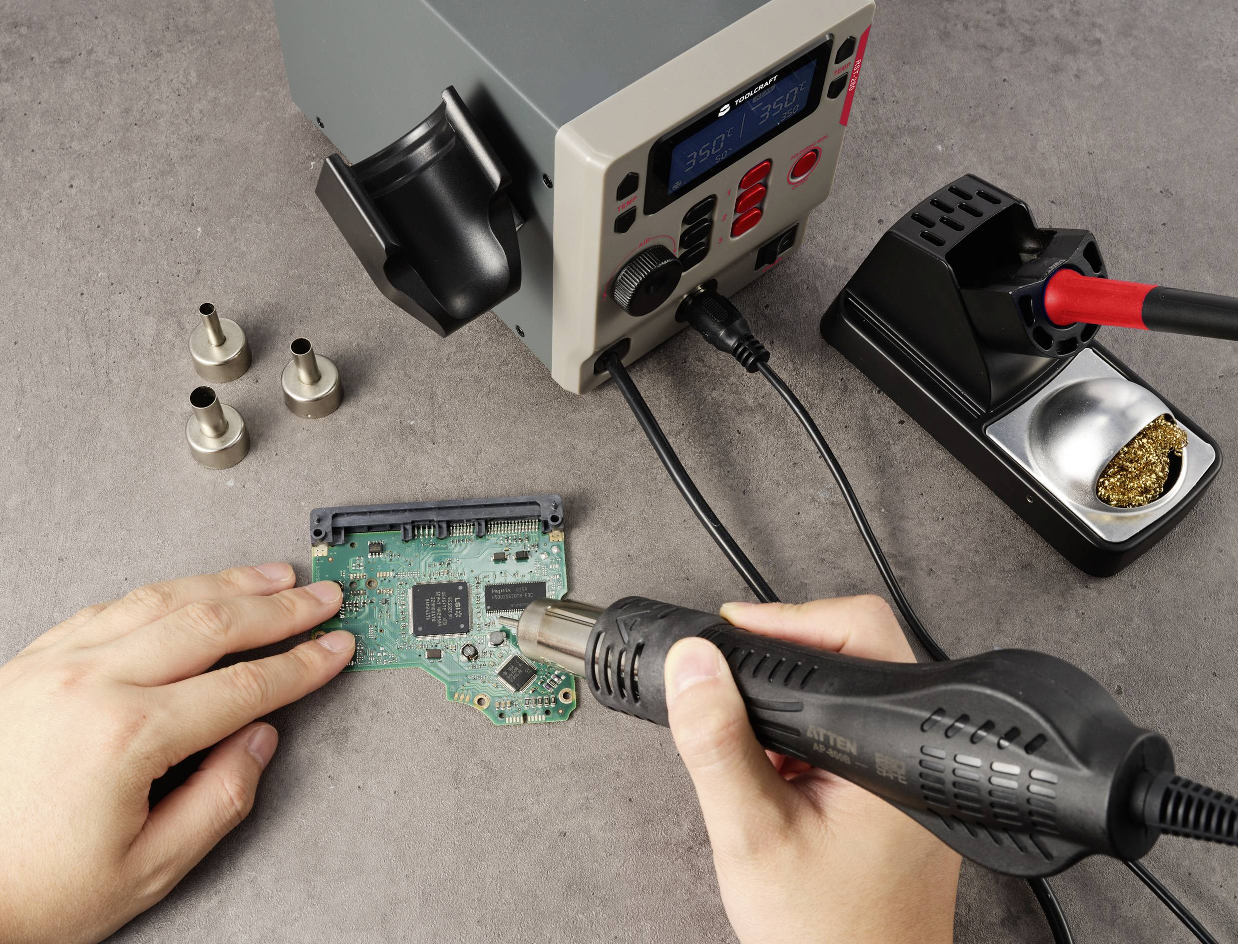 Person verwendet eine Heißluftpistole zum Löten auf einer Leiterplatte. Tools und Zubehör liegen auf dem Tisch neben der Platine.
