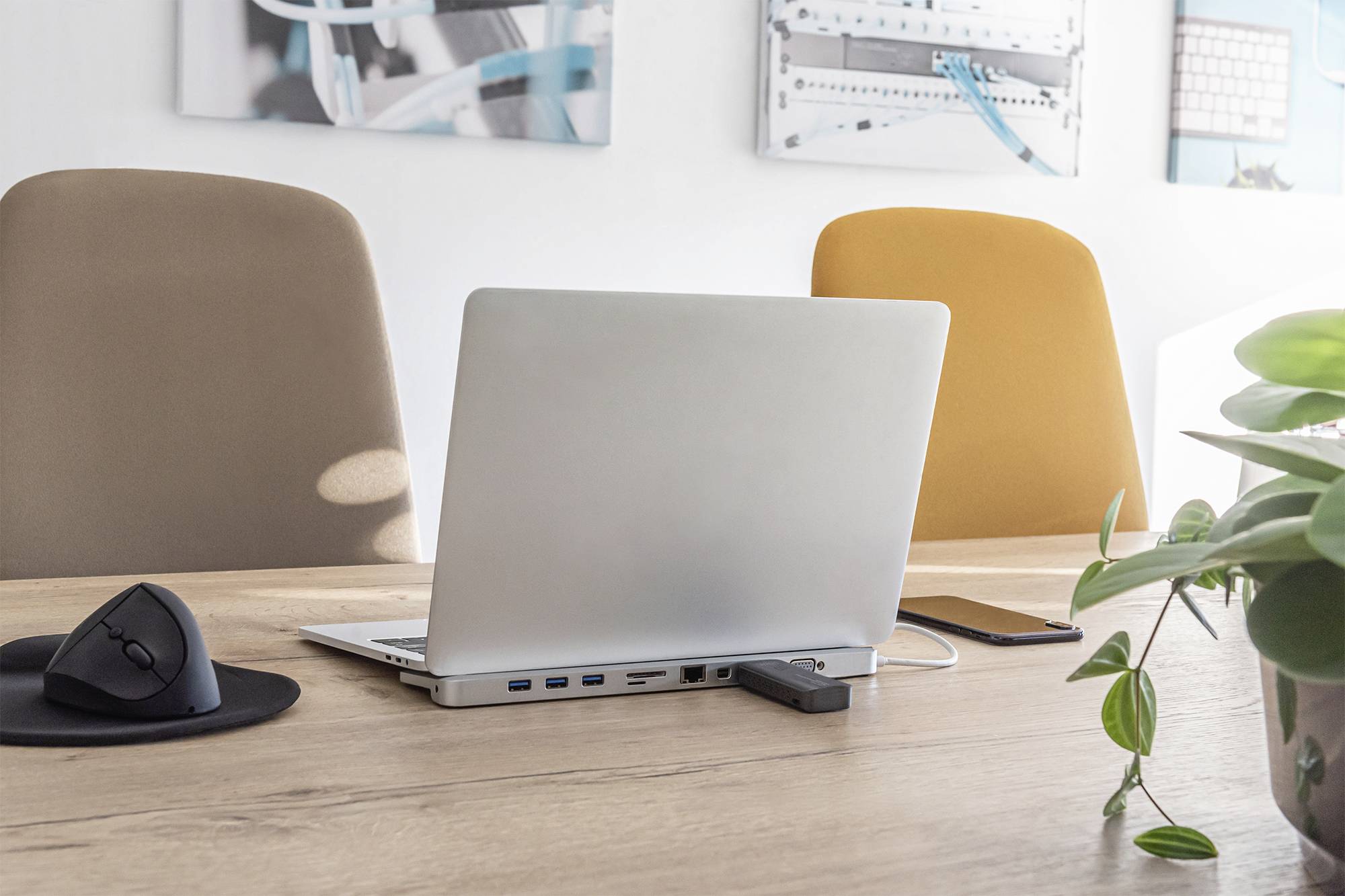 Ein Laptop steht auf einem Holzschreibtisch zwischen zwei leeren Stühlen. Daneben liegen eine Computermaus und ein Smartphone.