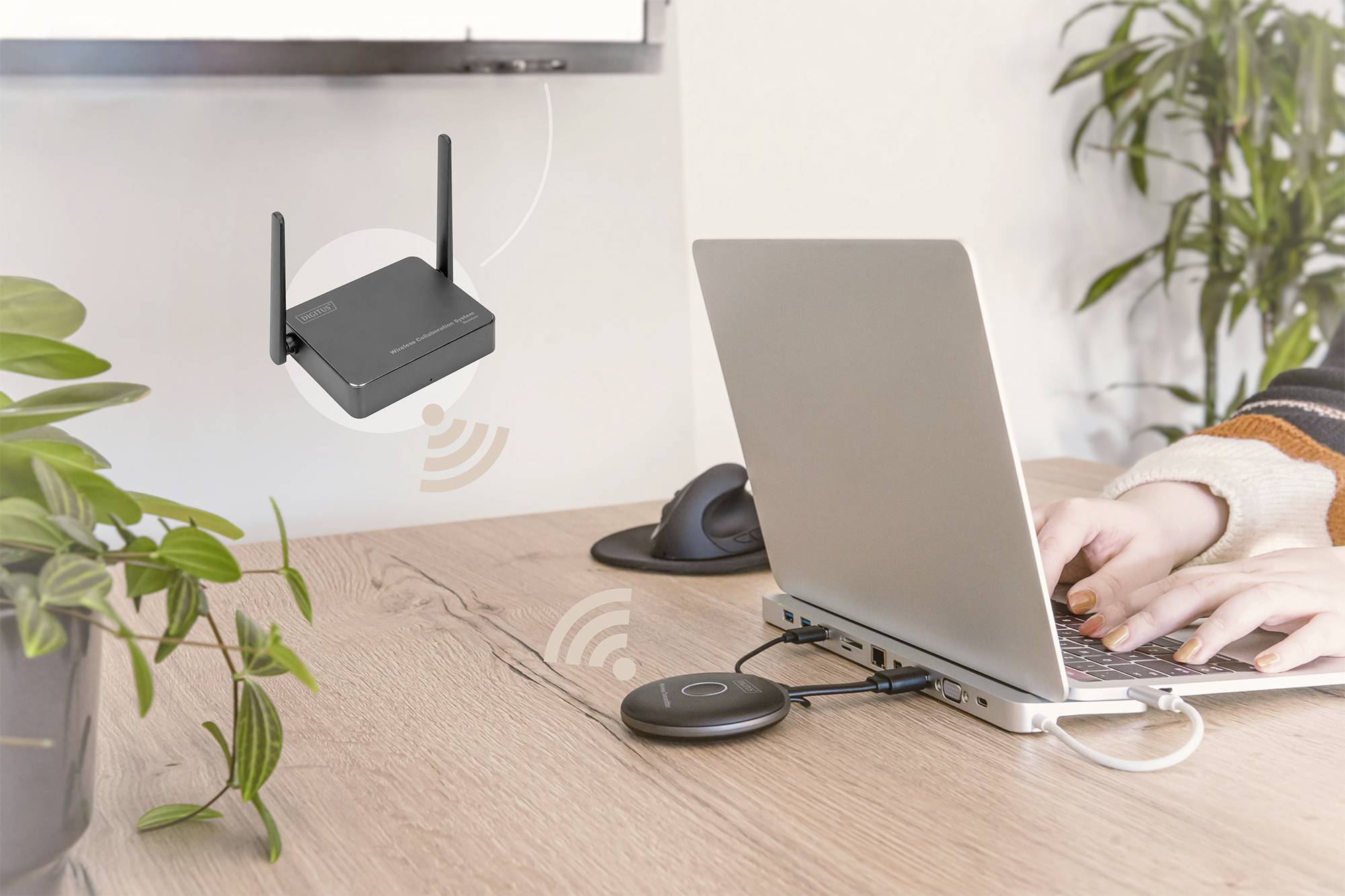 Eine Person tippt auf einem Laptop, der mit einem drahtlosen Adapter verbunden ist; im Hintergrund ein Router, der drahtlose Signale sendet.