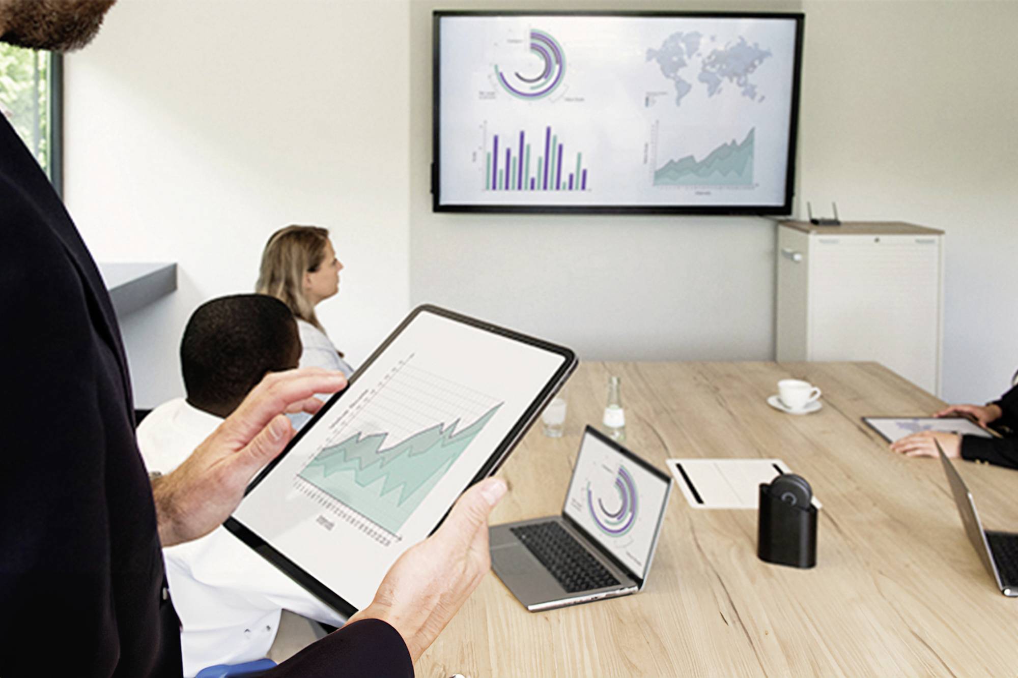 Personen in einem Konferenzraum betrachten auf Tabletts und Laptops Diagramme, während ein großer Bildschirm mit Grafiken angezeigt wird.