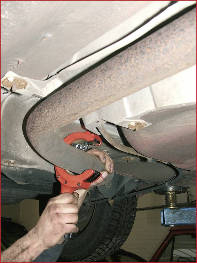 Eine Person repariert den Auspuff eines Autos in einer Werkstatt, hält ein Werkzeug, um den Auspuff unter dem Fahrzeug zu befestigen.