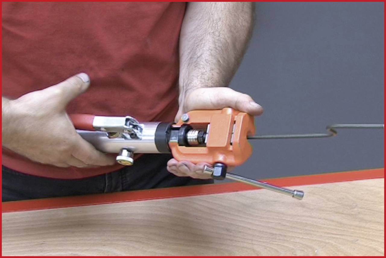 Eine Person hält ein rotes Handgerät mit einem langen Metallaufsatz, offenbar für eine Reparatur oder Montage über einem Holztisch.