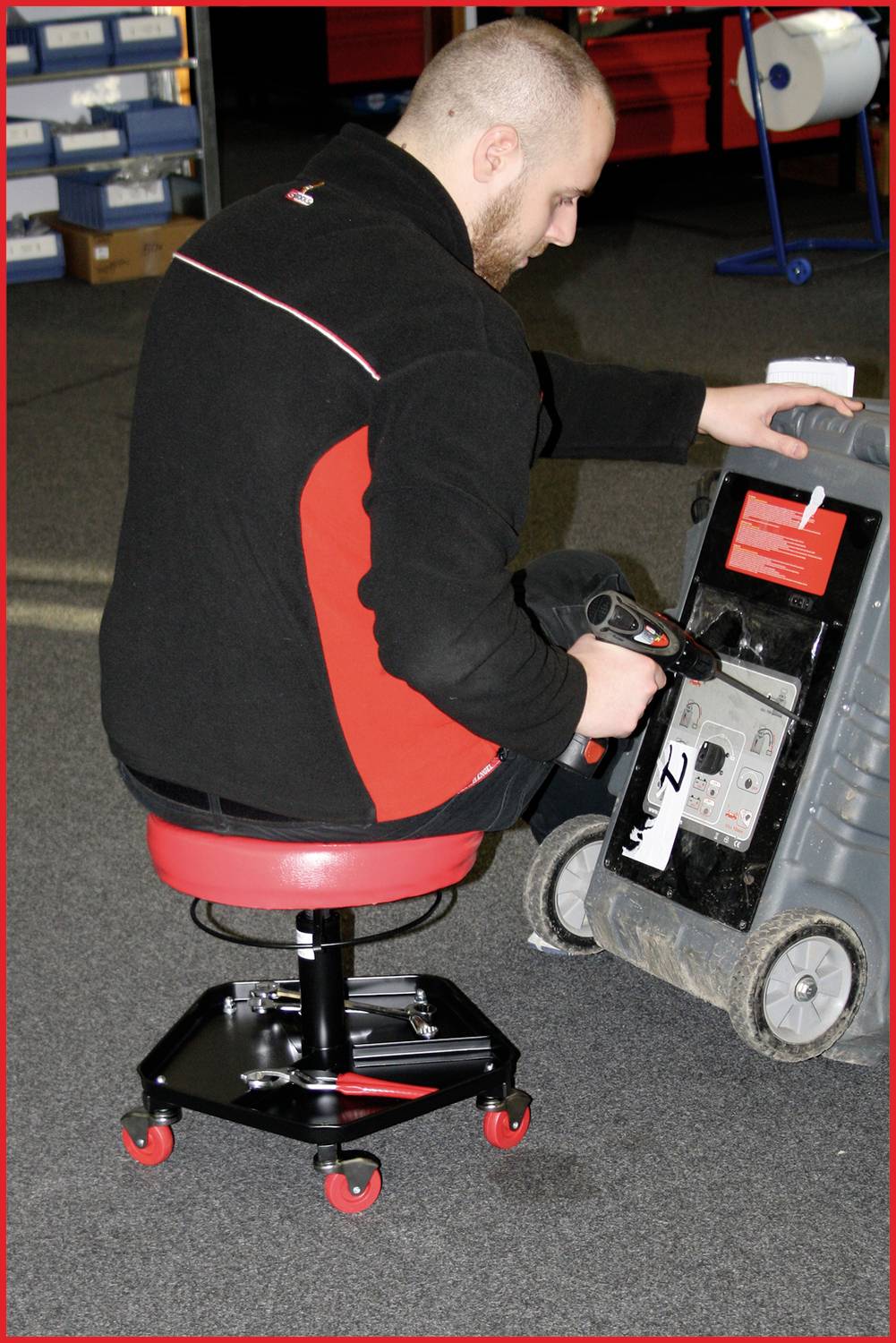 Ein Mann überprüft ein Gerät auf einem fahrbaren Sitz in einem Lager. Er hält ein Gerät und betrachtet die Vorderseite des größeren Objekts.