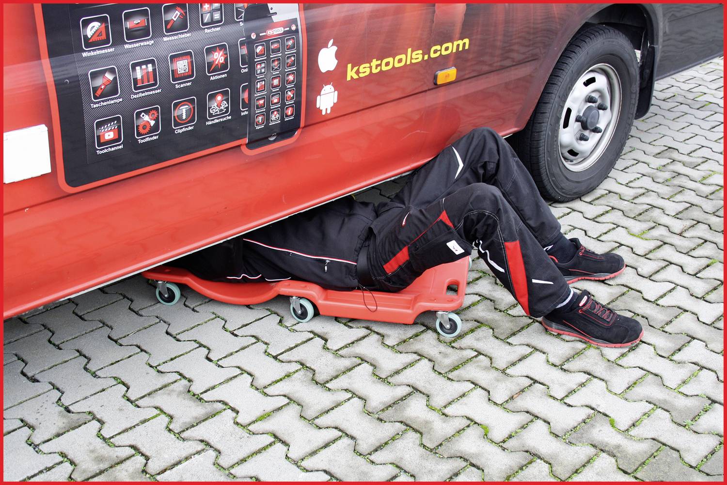 Ein Mechaniker arbeitet auf einem Rollbrett unter einem roten Lieferwagen auf einem gepflasterten Parkplatz.