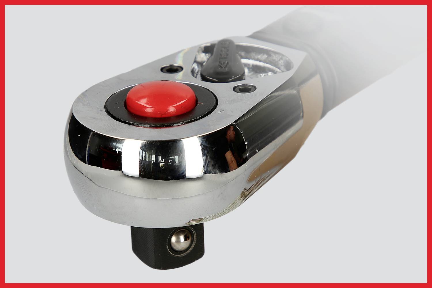 Nahaufnahme eines verchromten Werkzeugs mit rotem Knopf, möglicherweise ein Drehmomentschlüsselgriff, auf grauem Hintergrund.